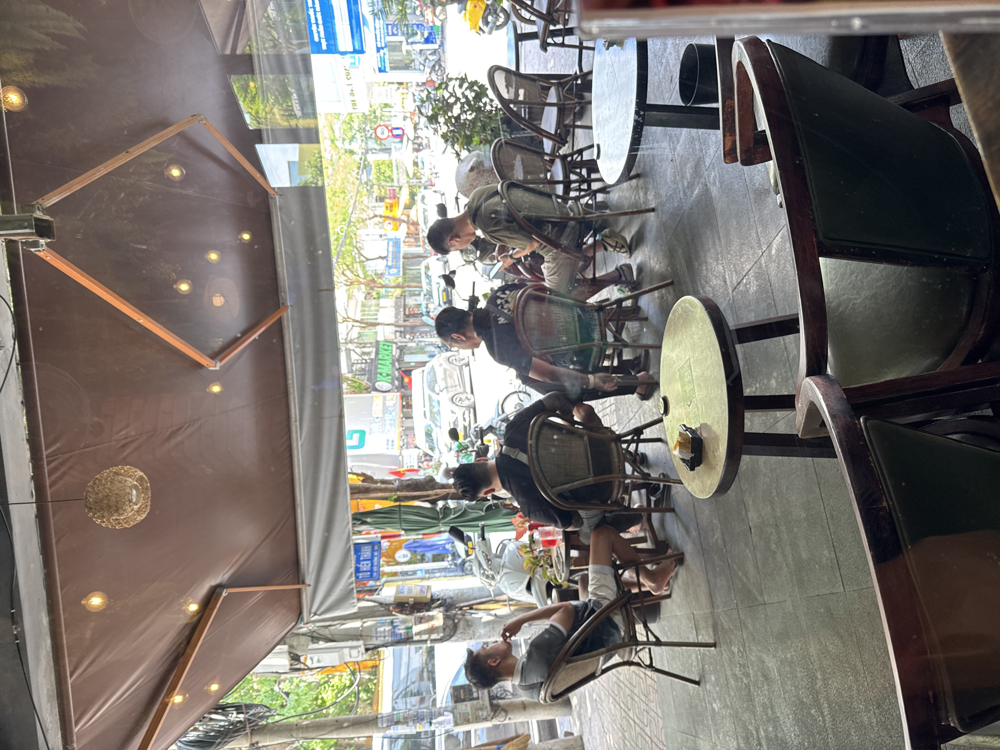 The image depicts an outdoor seating area of a cafe in Nha Trang, Vietnam. In the foreground, several people are seated at tables and chairs, engaged in conversation and enjoying their drinks. The cafe is covered by a brown awning with decorative lights, and the flooring is made of grey tiles. Outside the cafe, a street scene is visible with motorcycles, cars, and pedestrians. The atmosphere appears relaxed and casual, with people socializing and enjoying the pleasant weather.