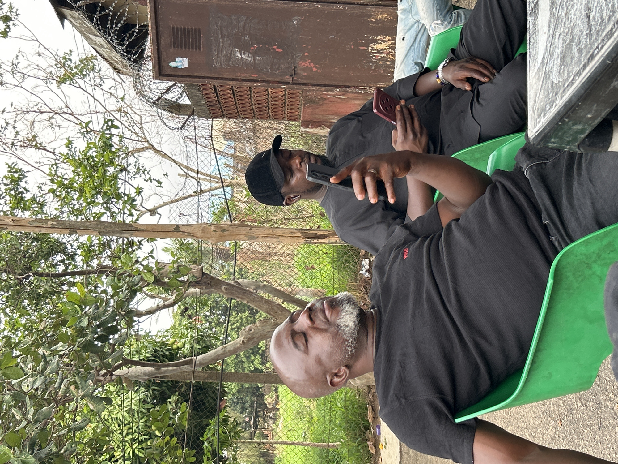 Two African men are sitting on green plastic chairs outdoors near a table, both looking at their phones. The first man, in the foreground, has a shaved head with graying facial hair and is wearing a black shirt with a red logo. The second man, positioned behind and to the left, is wearing a black shirt and a black baseball cap. He is also holding a phone. They are sitting near a wire fence and trees with green foliage. In the background, there is a rusty metal structure with barbed wire on top. The location is Jos, Nigeria.
