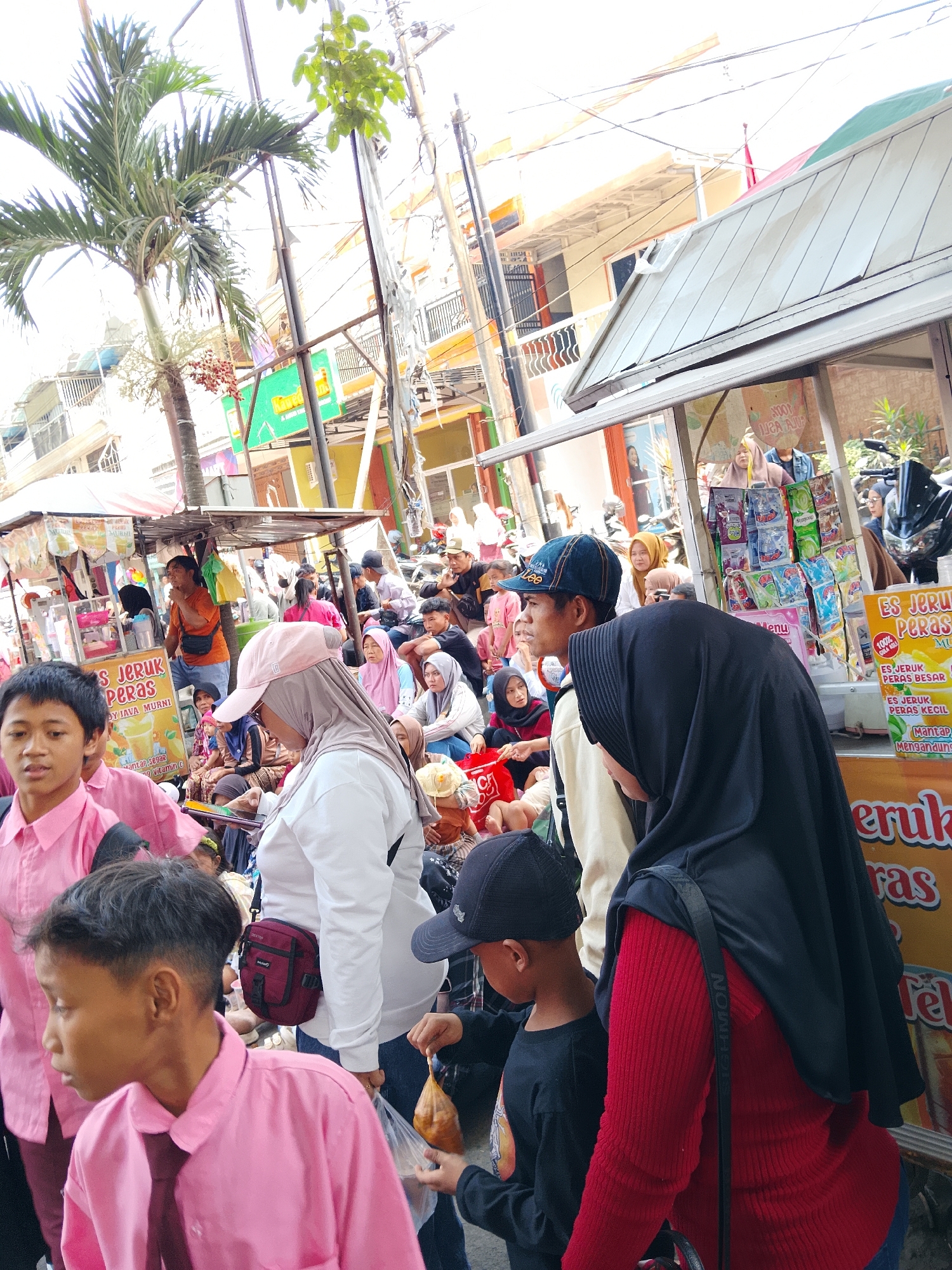 This is a vertical, eye-level, medium shot of a bustling street scene in Kertosono, Indonesia. The scene is dominated by a crowd of people gathered around a roadside stall selling drinks and snacks. In the foreground, a young boy in a pink collared shirt and tie looks to his left. To his right, a woman wearing a black hijab and a red knitted top is partially visible. Behind them, a woman in a pink hijab and white long-sleeved shirt looks down at her phone.
The stall displays various packaged drinks and snacks. In the background, buildings with colorful facades line the street, and overhead, a tangle of electrical wires crisscrosses the sky. A palm tree with drooping fronds is visible on the left side of the image. The overall impression is one of a lively, everyday street scene in a tropical urban environment.