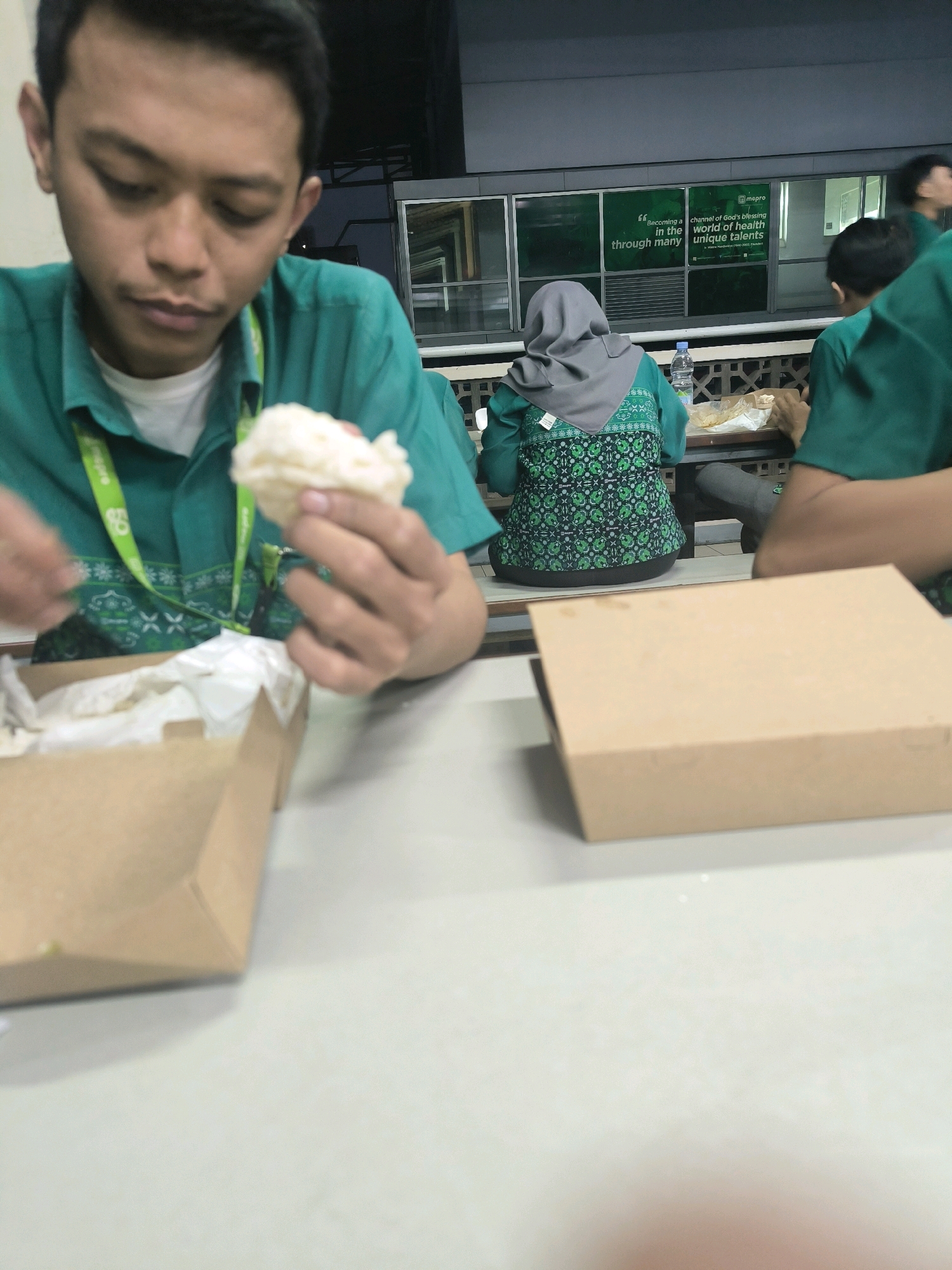 A close-up, slightly out-of-focus, medium shot captures a young man wearing a teal collared shirt with a white t-shirt underneath and a green lanyard. He is looking down at a piece of white food in his right hand, which he holds between his thumb and index finger. His left hand is partially visible, appearing to be reaching towards an open brown paper box in front of him, which contains white paper lining and some food.
In the background, a woman wearing a grey hijab and a teal shirt with a green and white patterned vest is seated at a table. She is facing away from the camera, and her hands are not clearly visible. Another person, also wearing a teal shirt, is seated to her right, facing away from the camera. A water bottle is visible on the table in front of the woman.
Behind the seated individuals, a windowed wall features a green sign with white text. The text is partially legible and includes the words "Becoming a channel of God's blessing" and "world of health unique talents." The wall appears to be part of a building's exterior, with some outdoor elements visible through the windows, including what looks like a metal grating and building structures.
The overall setting appears to be an indoor eating area with tables and chairs. The lighting is moderate, with some artificial light present. The image does not contain any specific visual cues that place it within the city of Bandung, Indonesia.