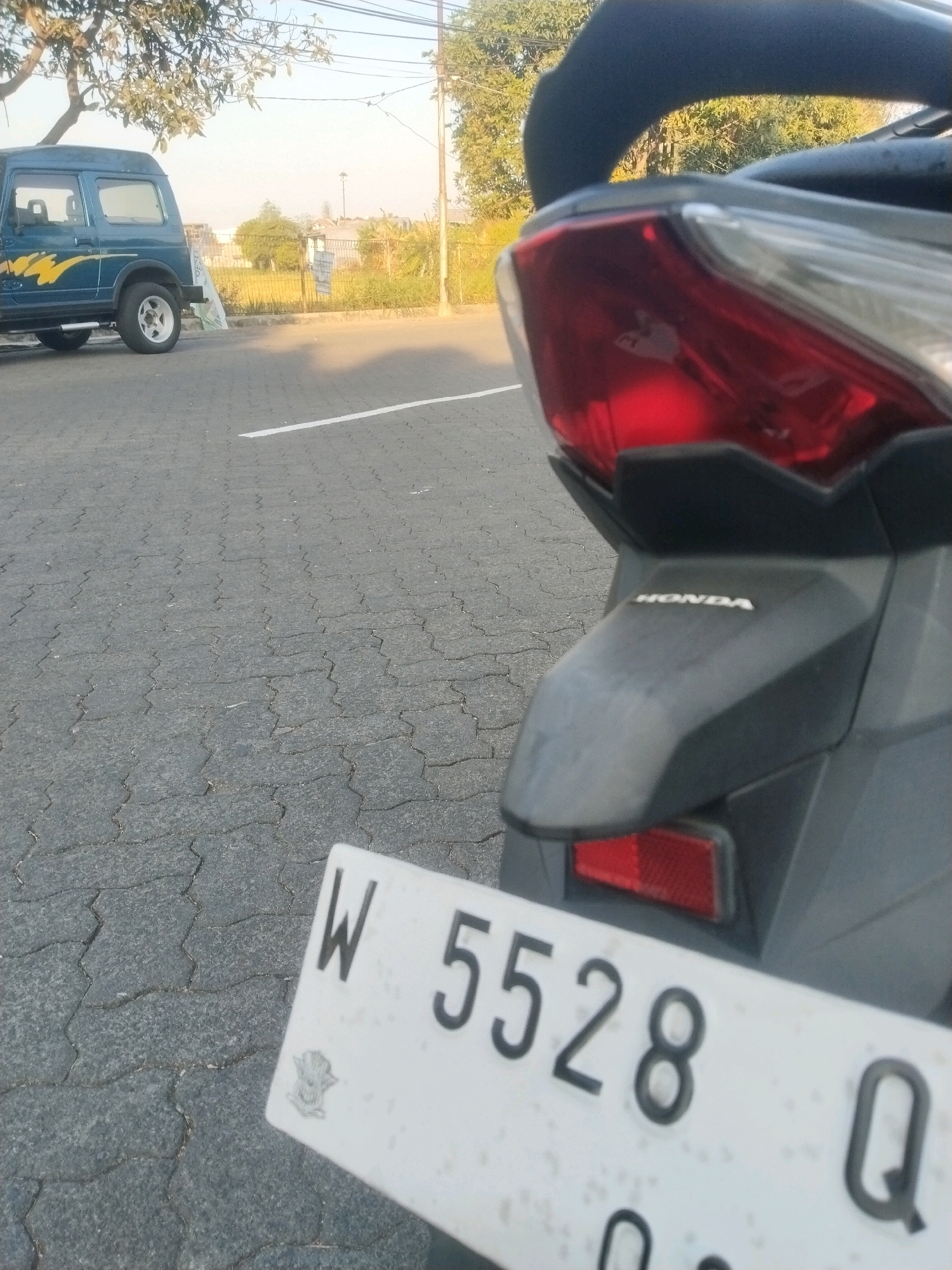 The image depicts a close-up view of a motorcycle's rear, focusing on its license plate and taillight assembly. The license plate, a white rectangle with black lettering, displays "W 5528 Q". Below the taillight, the "HONDA" logo is visible on the motorcycle's body. In the background, a blue off-road vehicle is parked to the left. The scene is set on an asphalt-paved area, possibly a parking lot, with a white line marking a boundary. The environment suggests an outdoor setting with trees and buildings visible in the distance, consistent with a location in Pepelegi, Indonesia. The lighting indicates it is daytime, likely late afternoon given the warm tones. There are no people visible in the frame.