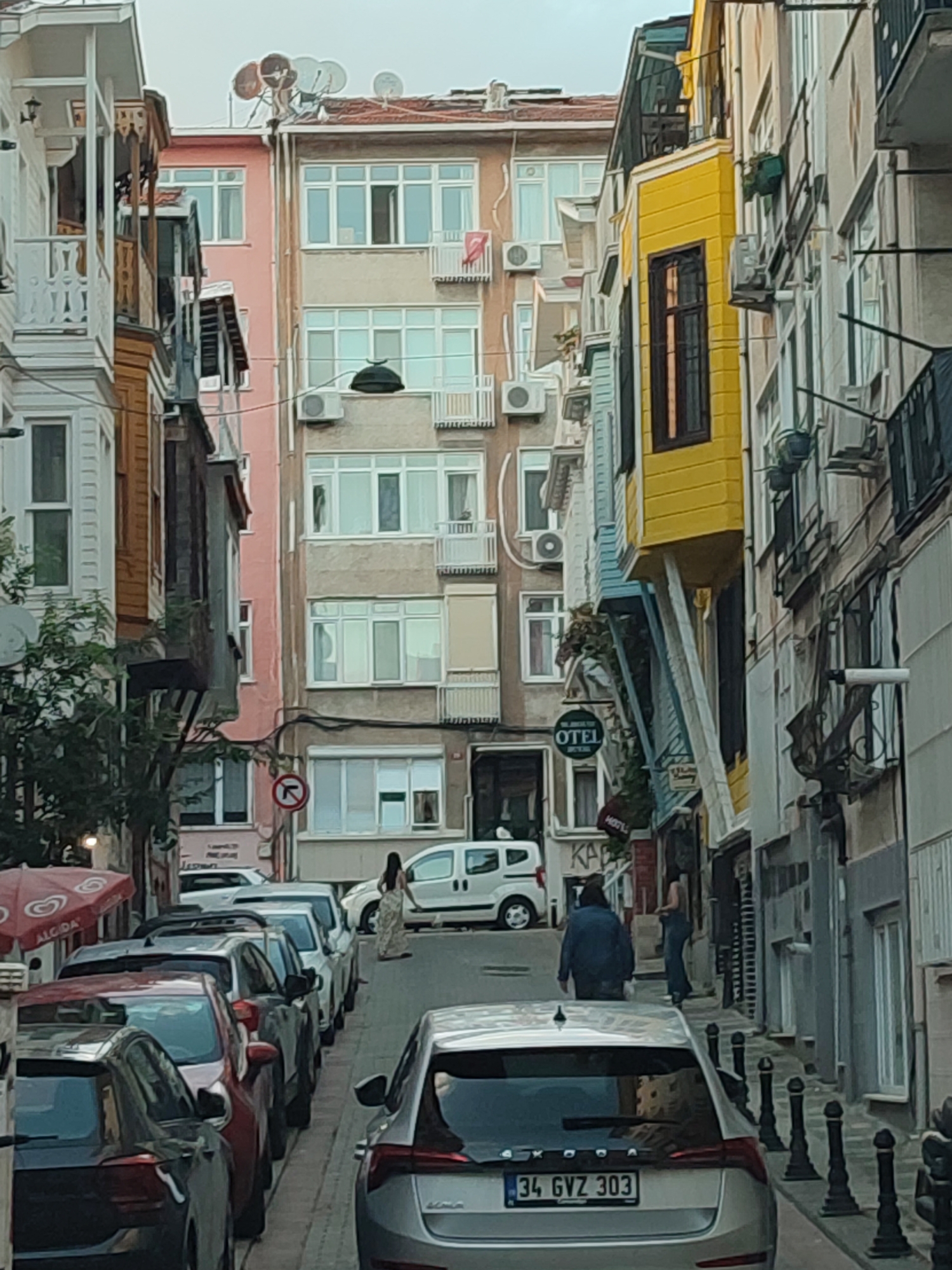 The image depicts a street scene in Kadıköy, Turkey, characterized by narrow, densely packed buildings of varying architectural styles and colors. The street is lined with parked cars, indicating it is a public thoroughfare. The buildings feature multiple stories, with windows and balconies visible. One building is painted bright yellow, contrasting with the predominantly pastel and neutral tones of its neighbors. Signs for "OTEL" are visible, suggesting the presence of accommodation establishments. People are present in the scene, walking along the street and interacting with the environment. The sky appears overcast, and the overall lighting suggests it is daytime. The presence of satellite dishes on rooftops is also notable. The street is paved, and bollards line one side, suggesting pedestrian and vehicle traffic management.