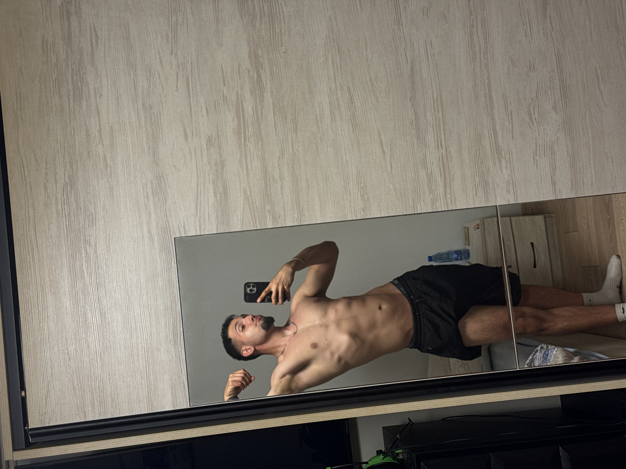 A young man with short dark hair and a goatee is standing in front of a mirrored wardrobe, taking a selfie. He is shirtless, wearing black shorts and white socks. He is flexing his arm and looking at his reflection. The wardrobe is light wood with vertical lines. In the reflection, a water bottle and a nightstand with drawers are visible. The floor appears to be wood. The image is a horizontal shot. The man's location in the city of Shumen, Bulgaria, is not determinable from the image.