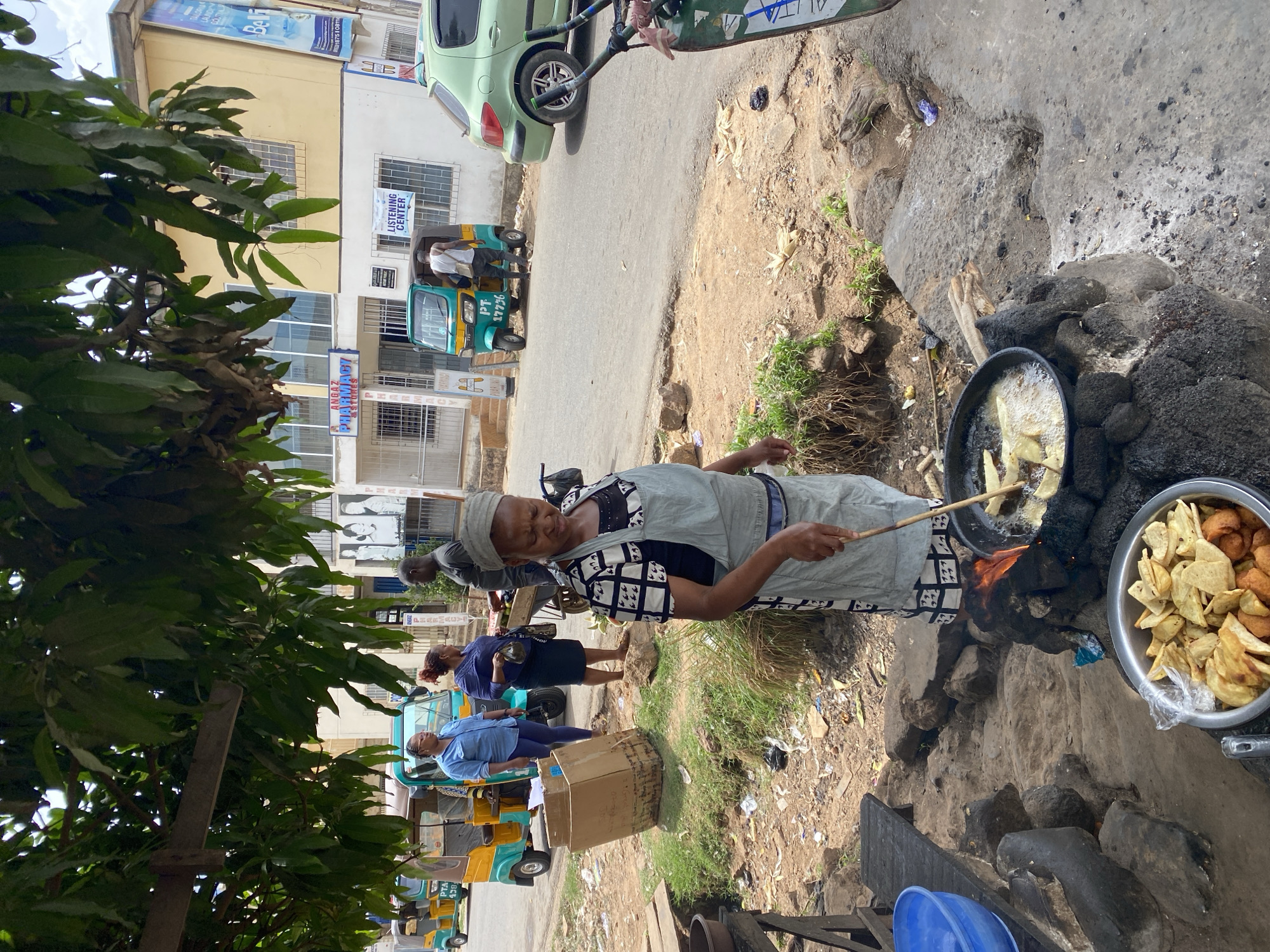 A woman fries food by the roadside in Jos, Nigeria. The woman, wearing a patterned head covering, is frying food in a large pan over a wood fire. A bowl of cooked food sits next to the pan. The setting is a roadside with greenery and trash. In the background, there are buildings, people, and motorized rickshaws. A car is parked nearby.