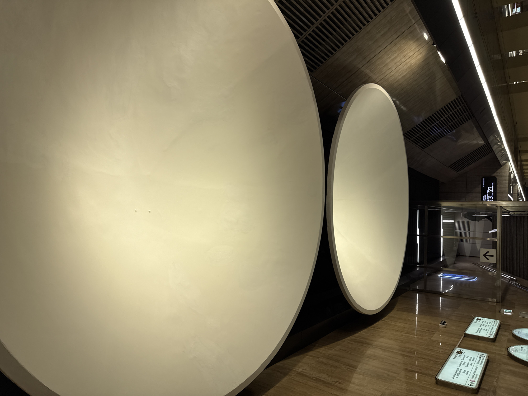 The image captures a scene within a Moscow Metro station, likely a newly constructed or renovated area. Two large, circular, white panels dominate the foreground, appearing to be artistic or architectural elements. The ground is covered with brown wood-like tiles.
In the background, there's a passage that appears to lead to another part of the station. The passage has a glass or transparent barrier with an arrow pointing to the left. Above the passage, a digital display shows the time as "13:21."
The ceiling is made of dark, grid-like material with some linear light fixtures running along its length. Small information plaques are visible on the floor near the right edge of the image.