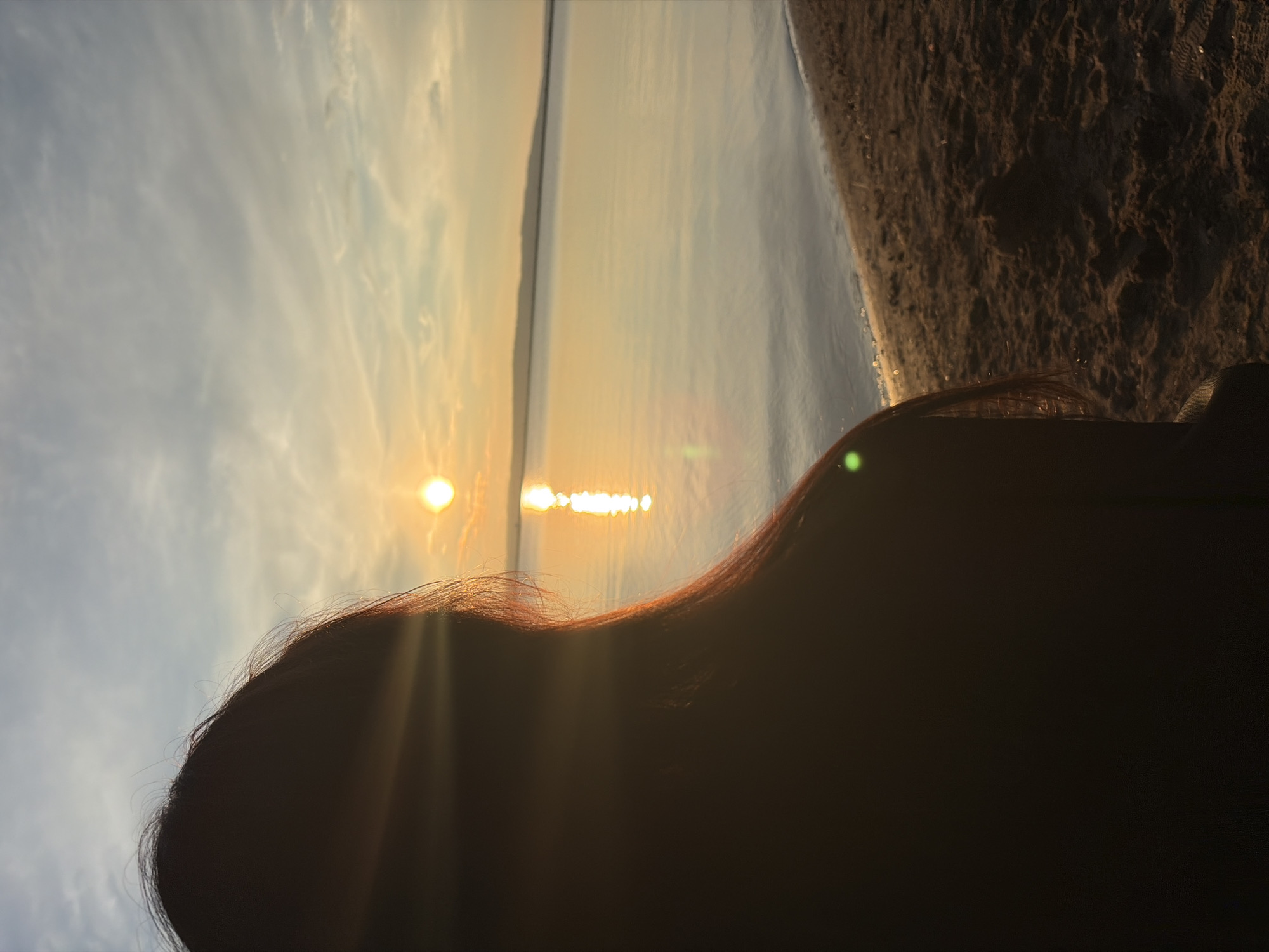 An over-the-shoulder shot shows a person's silhouette against a sunset over the Kama River in Chaikovsky, Russia. The sun, a bright orange orb, is low on the horizon, casting a shimmering reflection on the calm water. The sky above is a mix of blue and pale orange, with wispy clouds. The foreground features a sandy beach. The person's dark hair is visible, partially illuminated by the sun's rays. The overall scene evokes a peaceful, end-of-day atmosphere.