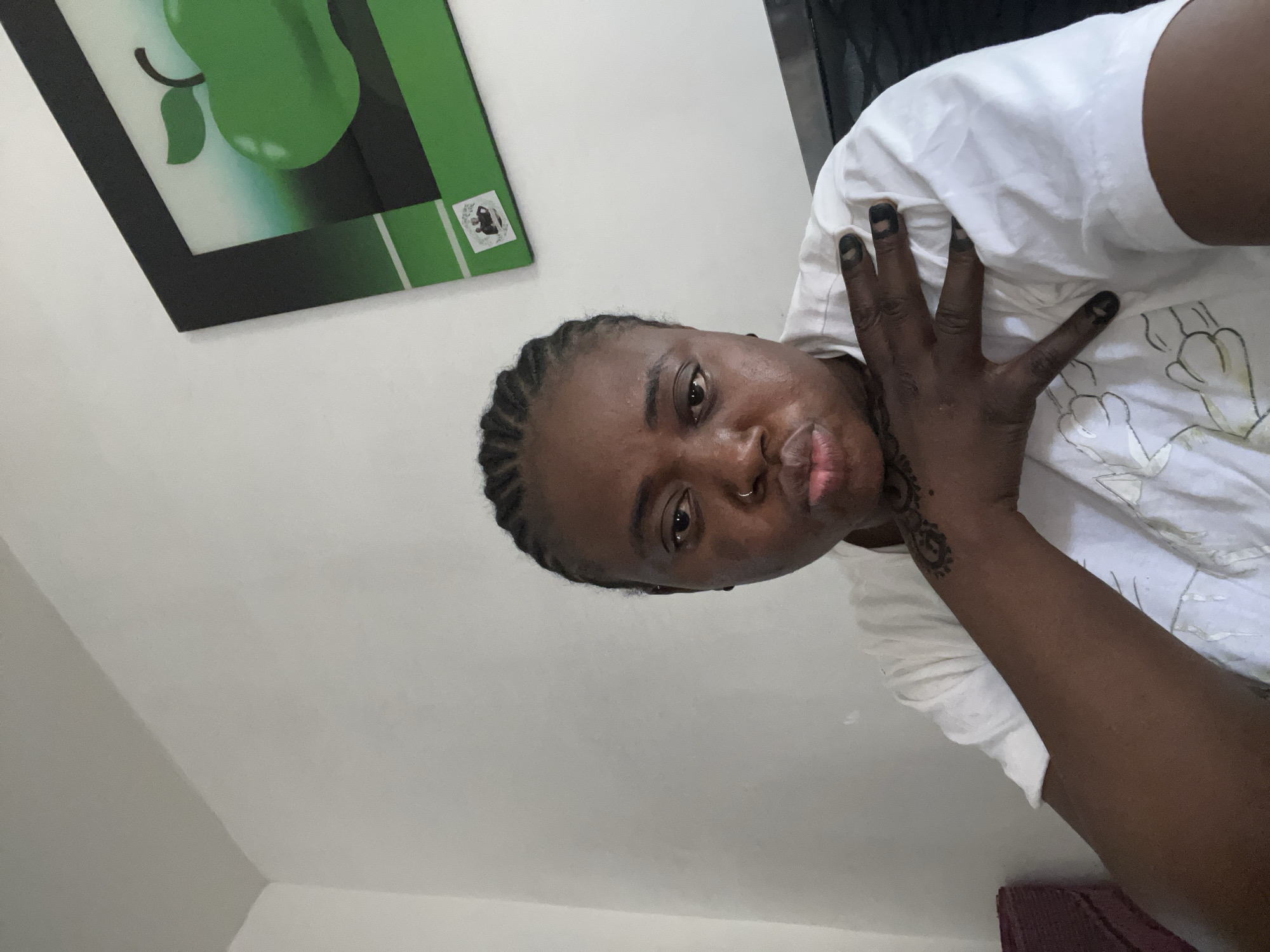A medium-shot, low-angle, slightly askew image shows a dark-skinned person with braided hair facing the camera. They are wearing a white shirt. Their left hand is raised, palm facing inward. There appears to be temporary tattoo or henna art on the forearm. The person's fingers appear to be discolored. The background consists of a light-colored wall. A framed image with a green pear design hangs on the wall in the upper left corner of the frame. The scene appears to be indoors, in a residential room in Jos, Nigeria. A corner of a burgundy-colored chair is visible at the bottom right.