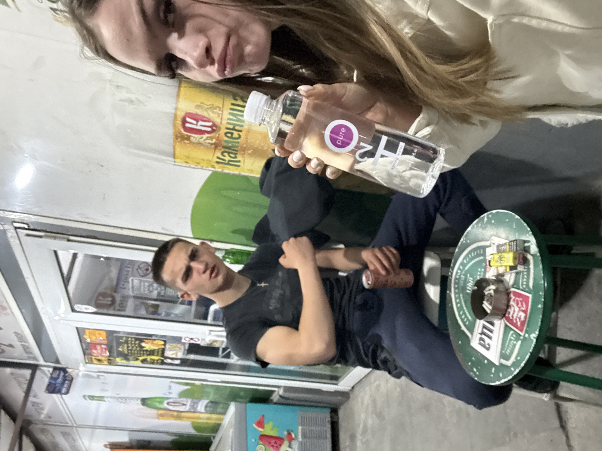 A medium shot of two people, a man and a woman, in front of a shop in Plovdiv, Bulgaria. The woman is holding a bottle of "Pure H2O Spring Water." The man is seated, holding a can. A small table is visible next to him with several items. The storefront has advertisements in the windows.