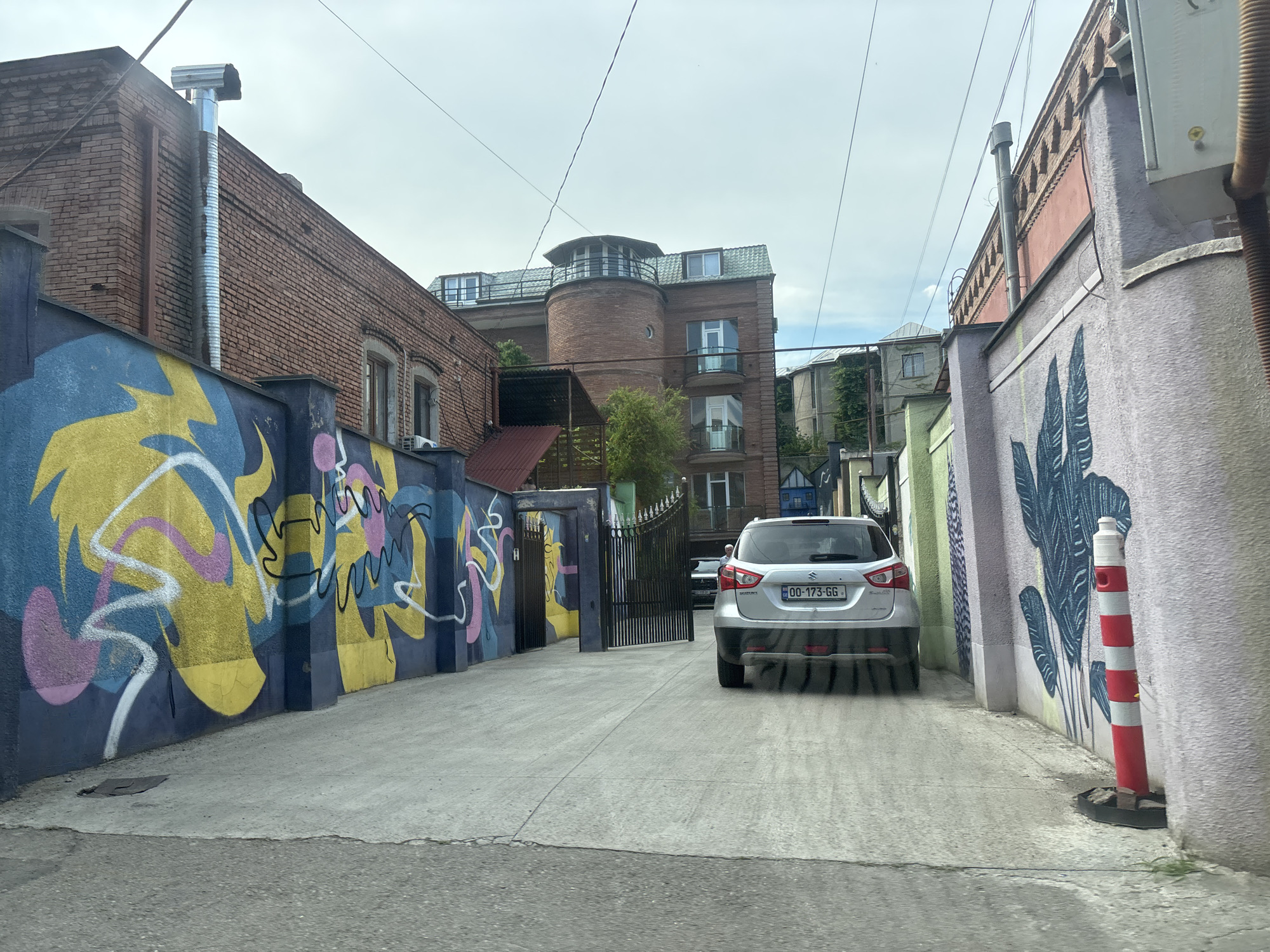 This is a medium-shot, eye-level photograph taken from a vehicle traveling down a narrow, paved alleyway in Tbilisi, Georgia. The alley is flanked by buildings and walls adorned with colorful graffiti.
On the left, a brick building with a metal pipe extending from its roof is visible, with a wall covered in abstract, vibrant graffiti featuring yellow, blue, pink, and white shapes. To the right, a pastel-colored wall is decorated with a large, stylized illustration of dark blue leaves. A red and white striped bollard stands in front of this wall.
In the background, a multi-story brick building with a cylindrical turret and rounded windows is prominent. Another building with green roofing is partially visible. A silver Suzuki SUV is parked in the alley, facing away from the camera. Its license plate reads "00-173-GG." A black car is partially visible through a metal gate further down the alley.
The sky is overcast, suggesting a cloudy or possibly rainy day. The overall scene depicts an urban environment with artistic street art and a residential or mixed-use area. There are no people visibly present in the immediate vicinity of the vehicles. The time of day is indeterminate due to the overcast sky.
