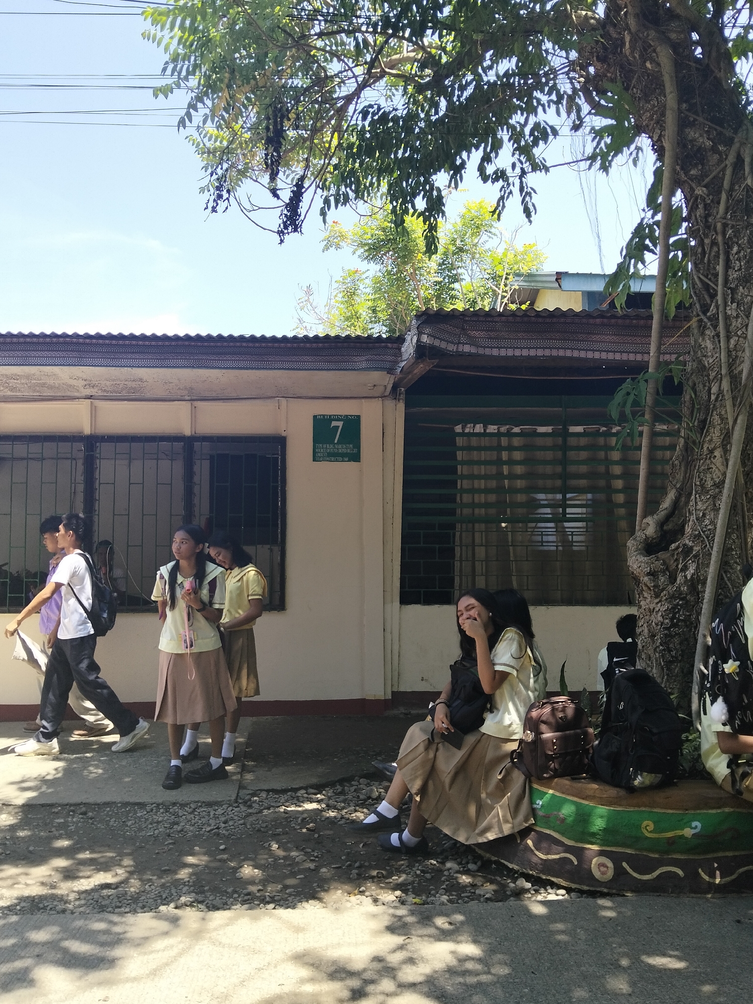 The image depicts a street scene outside a building identified as "BUILDING NO. 7" in Davao City, Philippines. A sign indicates the building type as "SOURCE OF FUNDS DEPARTMENT." Several students in school uniforms are visible. On the left, two male students walk past a building with barred windows, one carrying a backpack. In the center, two female students in similar uniforms are standing, one appearing to adjust her hair or clothing, and the other looking to the side. To the right, a female student is seated on a decorative bench, holding a phone and looking downwards. Another female student is partially visible to her right. The environment includes a building with a corrugated roof, a large tree with vines, and dappled sunlight on the paved ground. The overall atmosphere suggests a school or administrative building on a bright, sunny day.