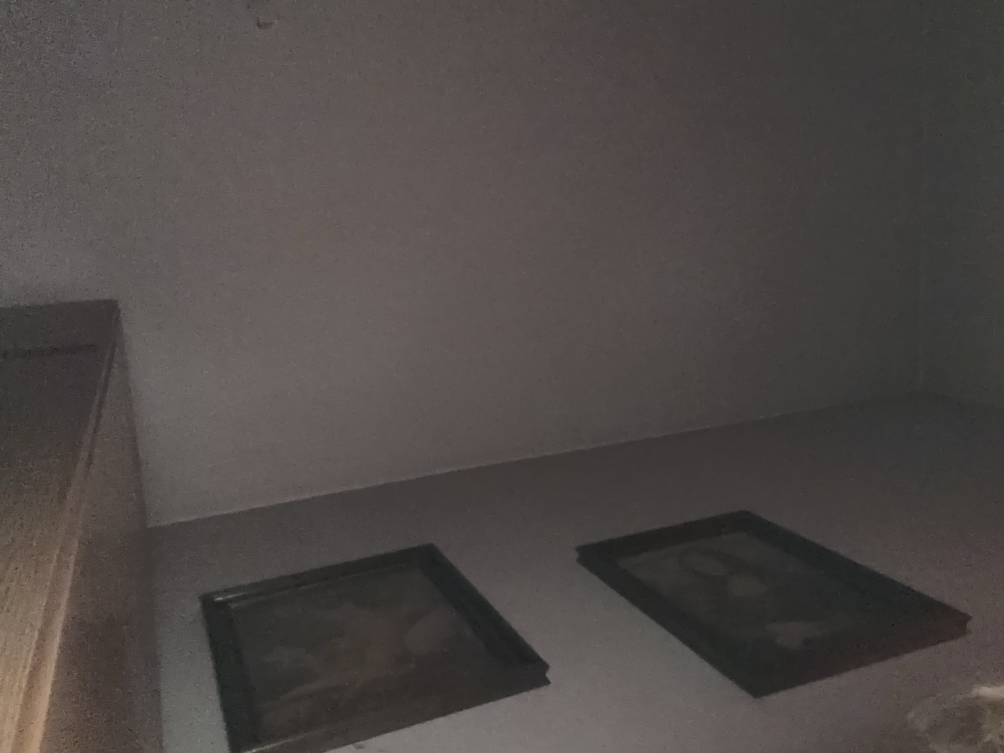 The image shows a corner of a room. There is a wooden cabinet on the left, and two framed pictures are on the floor. The room's walls and ceiling are a light, uniform color. The location is not discernible from the image.