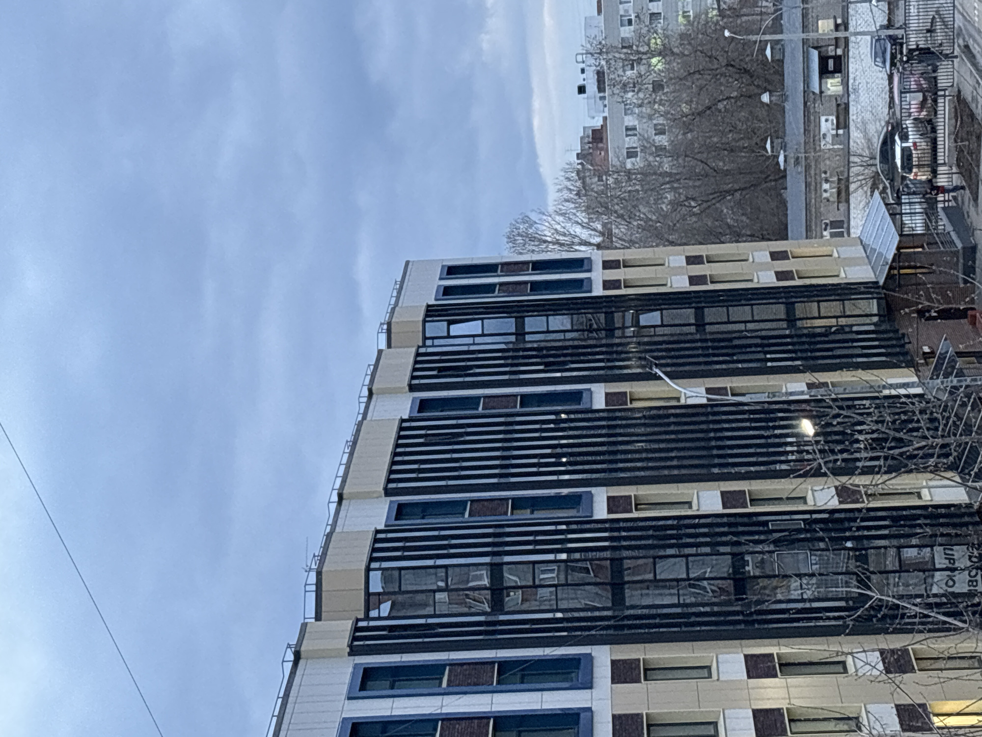 The image shows a multi-story building in Perm, Russia. The building has a modern design with a facade consisting of alternating rows of dark-framed windows and lighter-colored panels. There are dark horizontal structures extending out from the building, possibly balconies or sun shades. In the background, there is another building and bare trees, indicating it is likely autumn or winter. The sky is overcast. In the immediate foreground, there is a bare tree branch. There are parked cars visible in the background.