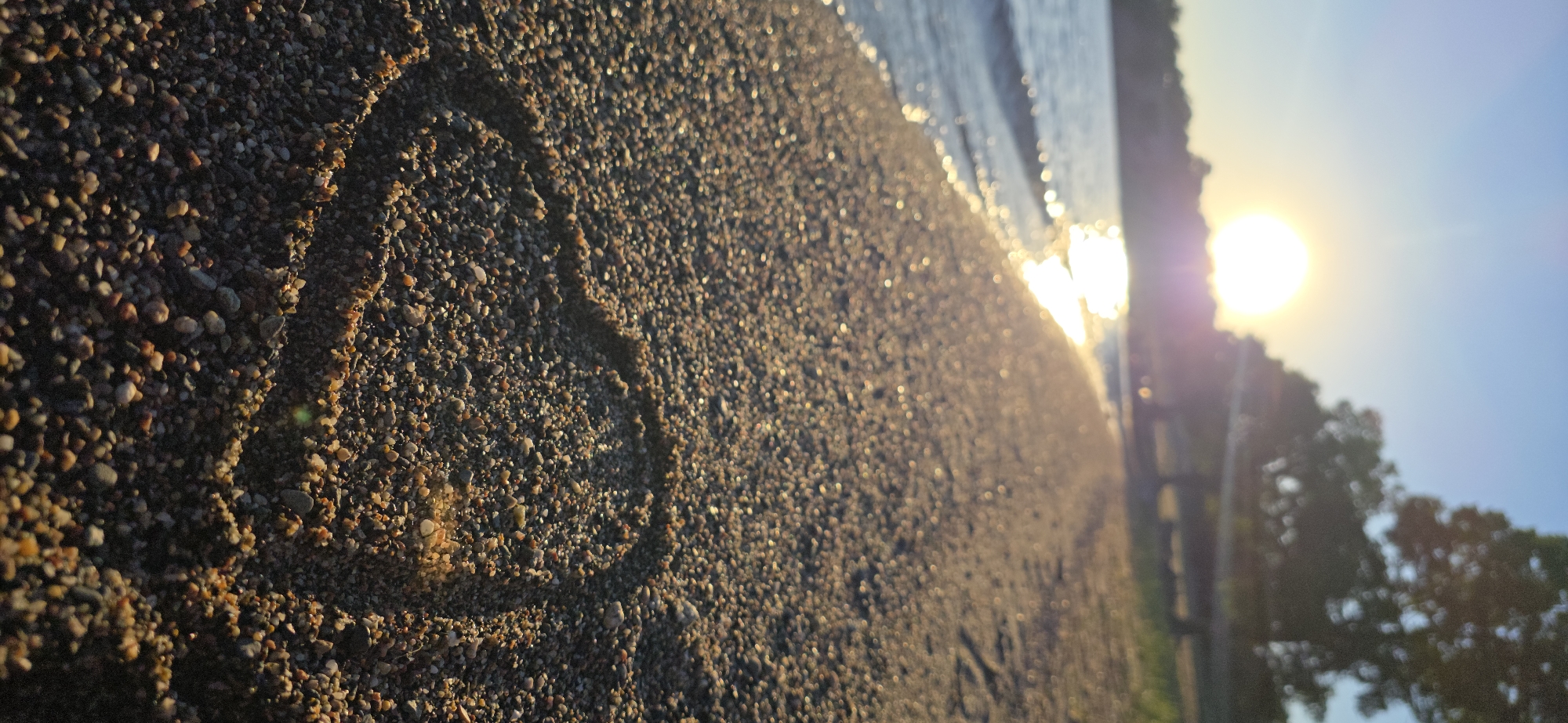A close-up, low-angle shot of a pebble-strewn beach on a sunny day. The sun, low in the sky, casts a warm glow on the scene, with its reflection shimmering on the gentle waves of a lake. In the foreground, a heart shape is drawn in the pebbles. The background shows a line of trees and a blurred horizon. The scene is peaceful and evokes a sense of romance. This image was taken in Issyk-Kul, Kyrgyzstan.