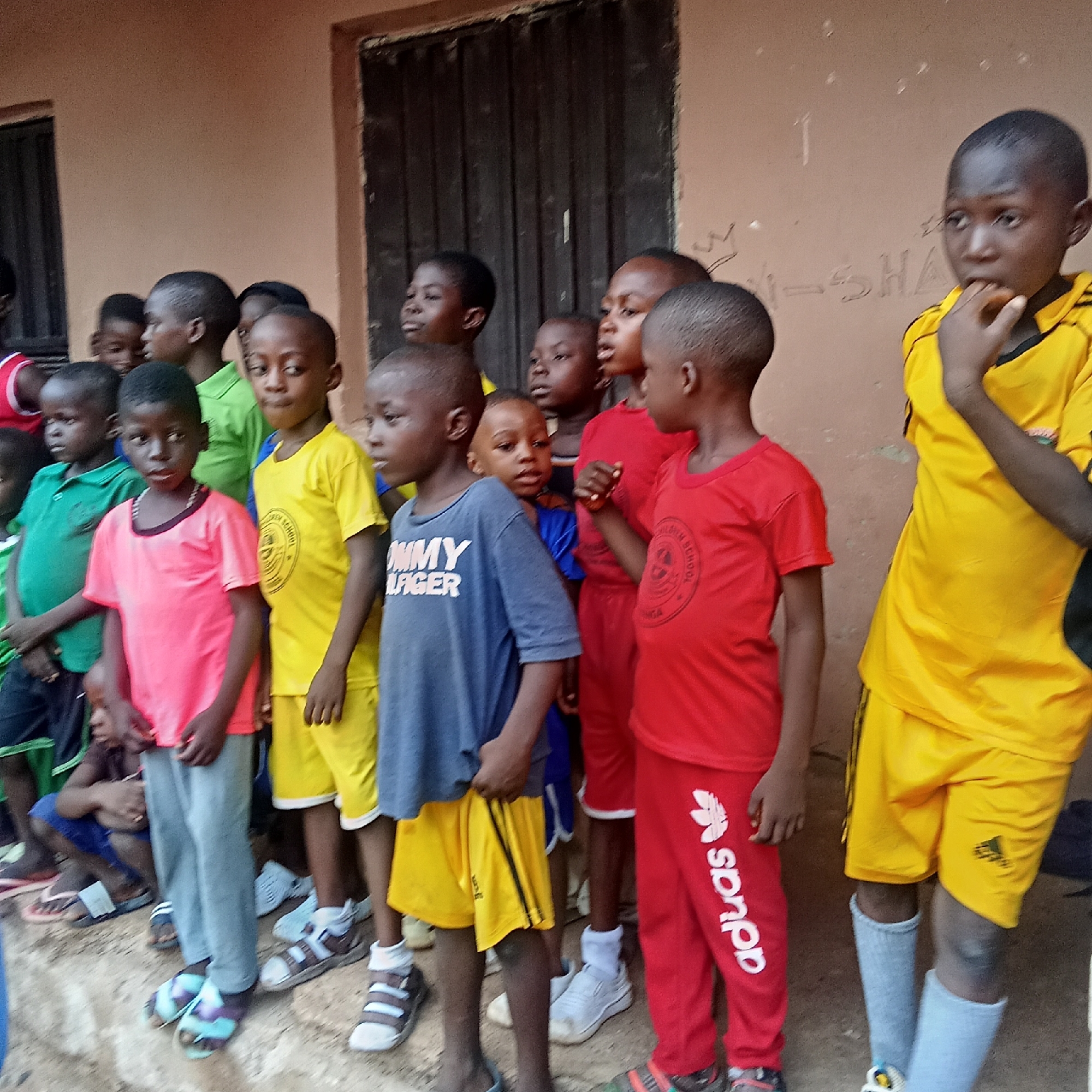 A group of children stand outdoors against a beige wall in what appears to be Akwanga, Nigeria. The wall has a dark door with vertical bars. The children are dressed in a variety of casual clothing, including t-shirts, shorts, and pants, some with logos. The clothing colors vary from red and yellow to pink, blue, and green. Some children wear sandals, while others wear sneakers or go barefoot. One child in a yellow Adidas outfit has his fingers in his mouth. The overall setting seems ordinary, and the children appear to be waiting or gathered together.