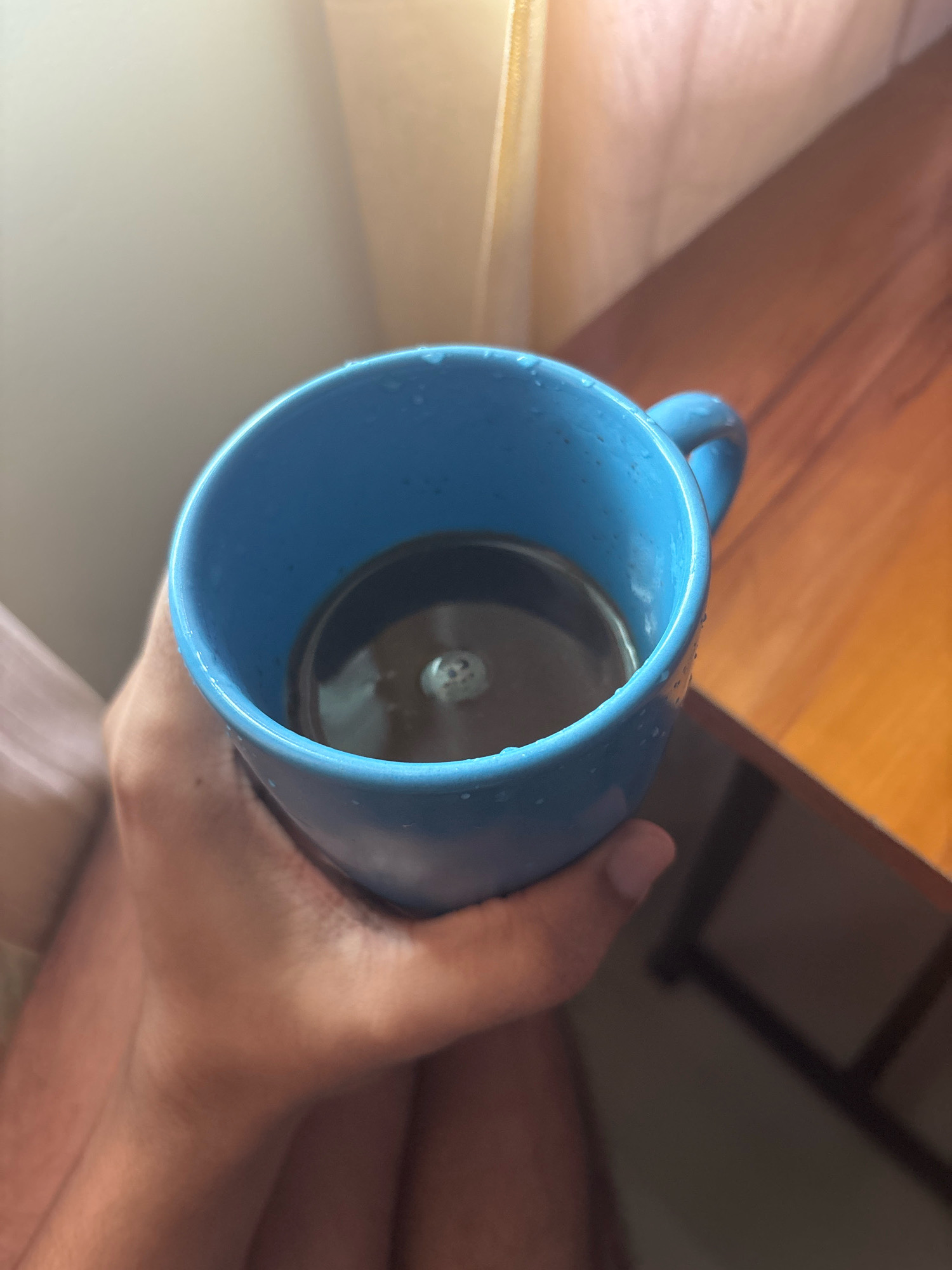 A close-up shot shows a hand holding a blue ceramic mug filled with a dark liquid, likely coffee, with a faint reflection on the surface. The mug has a handle visible on the right side. The background is softly blurred, with a wooden surface and a curtain in soft light. There is no discernible information about the location in the city of Bhatta, India.