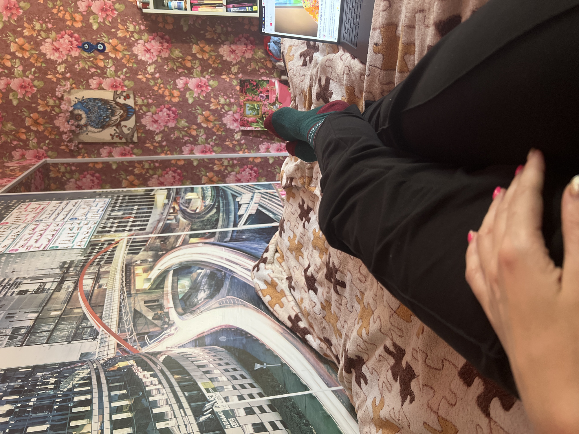 The image shows a person's legs resting on a patterned blanket. The person is wearing dark pants and green and red socks. A laptop is open on the blanket, displaying a web page with Russian text. In the background, a wall is covered with floral wallpaper, and a framed picture of an owl is hanging on it. There are also shelves with books visible at the top of the frame. To the left, a large framed image of a futuristic cityscape with curved roads and buildings is mounted on the wall, extending from floor to ceiling. A hand with pink nail polish is visible reaching towards the right side of the frame. The setting appears to be indoors, likely a bedroom or living room, with the presence of a bed or sofa suggested by the blanket and the relaxed posture of the person. The time of day is not discernible. The text on the laptop screen appears to be an article or blog post, with the phrase "КАК СТАТЬ ТОЙ, РАДИ КОГО УБИВ" visible, which translates to "How to become the one for whom they kill." Other legible text on the cityscape image includes "AAER," "CBRE," and "PLAT AURENOVE." The overall mood seems relaxed and casual, with the person engrossed in using their laptop.