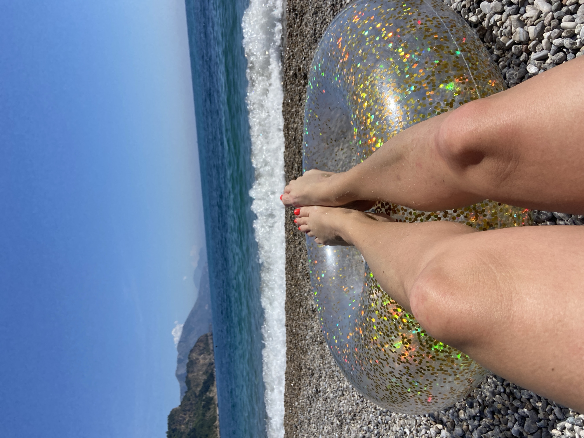 The image depicts a serene beach scene, captured from a first-person perspective, looking down towards the sea. In the immediate foreground, a person's tanned legs and feet are prominently displayed, with vibrant red nail polish on the toes. The feet are resting on a transparent inflatable ring, which is filled with shimmering golden glitter. This ring and the person's legs are situated on a pebble beach, composed of various shades of grey and brown stones.
Beyond the pebbly shore, white-crested waves gently break as they meet the land, indicating the clear blue sea. The calm, expansive blue water stretches into the distance, where rugged, dark green mountains rise dramatically from the coastline. The sky above is a brilliant, cloudless blue, suggesting bright, sunny weather during the day. The scene conveys a sense of relaxation and leisure, likely a vacation in the coastal region of Prijevor, Montenegro, given the mountainous backdrop and clear waters. No text is visible in the image.