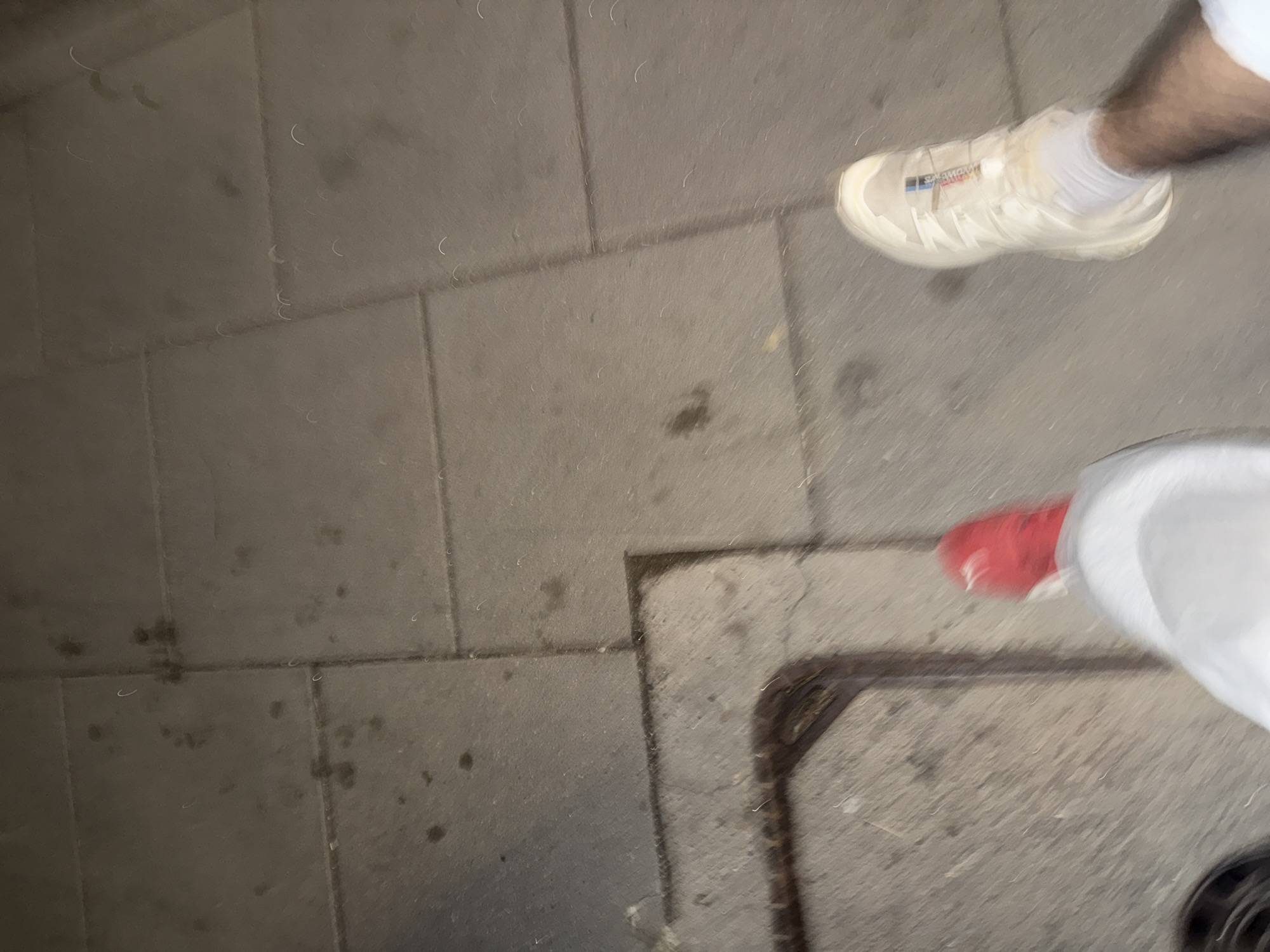 In this overhead shot, two pairs of feet are visible on a paved surface. One pair is adorned with bright red sneakers, while the other wears cream-colored athletic shoes with white socks. The ground beneath them is a mosaic of large, square concrete tiles, some of which are marked with dark, irregular stains. A metal grate or drain cover is situated in the lower left quadrant of the image, set into the pavement. The overall scene is one of a pedestrian walkway in an urban environment, possibly outdoors given the texture and condition of the paving stones.