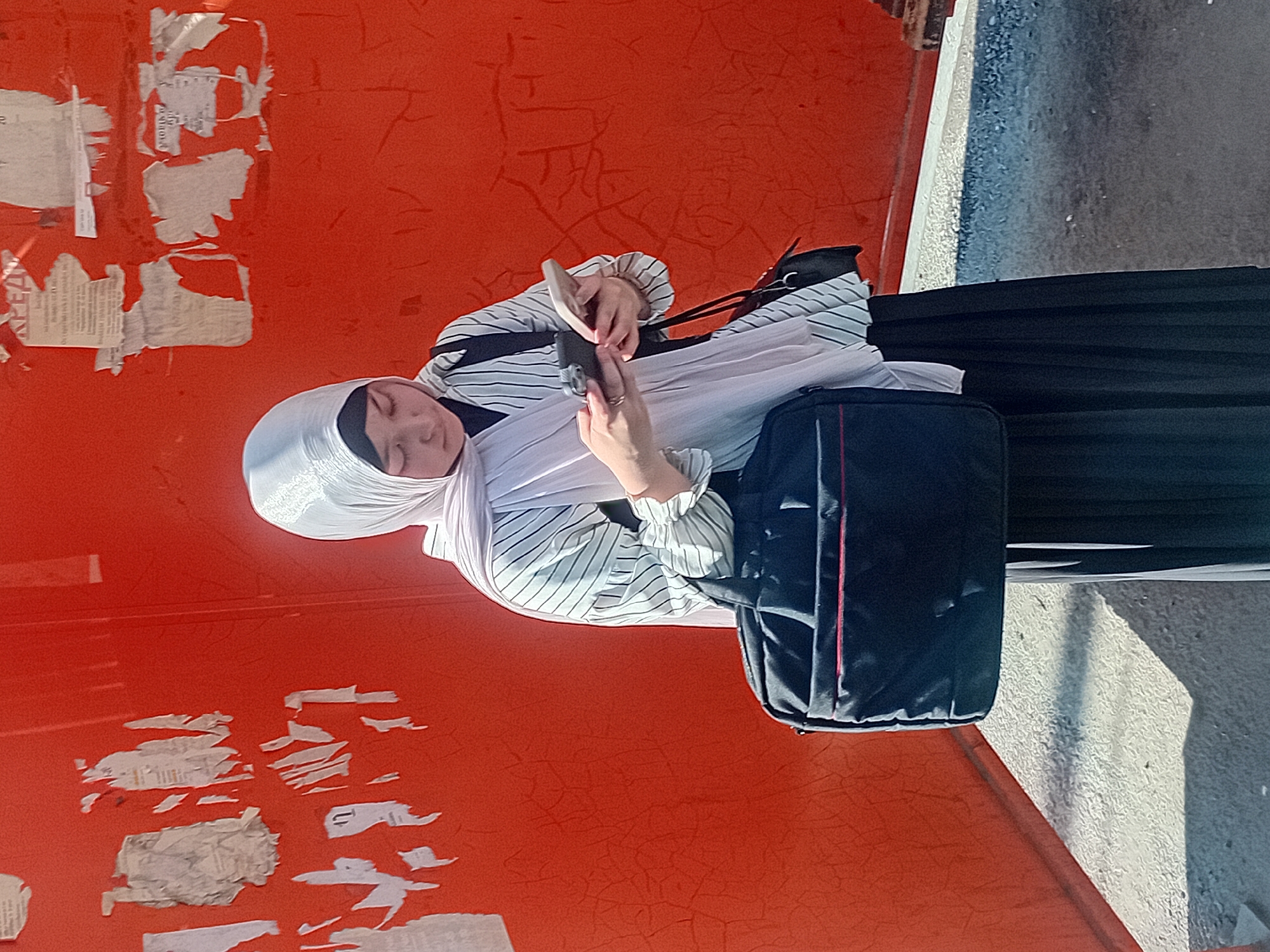 A young woman wearing a light-colored hijab and a striped shirt stands against a vibrant orange wall covered in torn posters. She is looking down at her mobile phones, one in each hand, and appears to be texting or browsing. A black messenger bag with red trim hangs from her left shoulder, and a smaller dark bag is slung over her right shoulder. The woman is also wearing a long, pleated black skirt. The ground beneath her is a mix of rough, textured pavement and shadow. The orange wall has a textured, cracked surface, and several pieces of paper are stuck to it, some torn and peeling away. The lighting suggests it is daytime and the sun is casting a shadow. The background hints at an outdoor urban environment, potentially a bus stop or a public wall in the city of Semey, Kazakhstan.