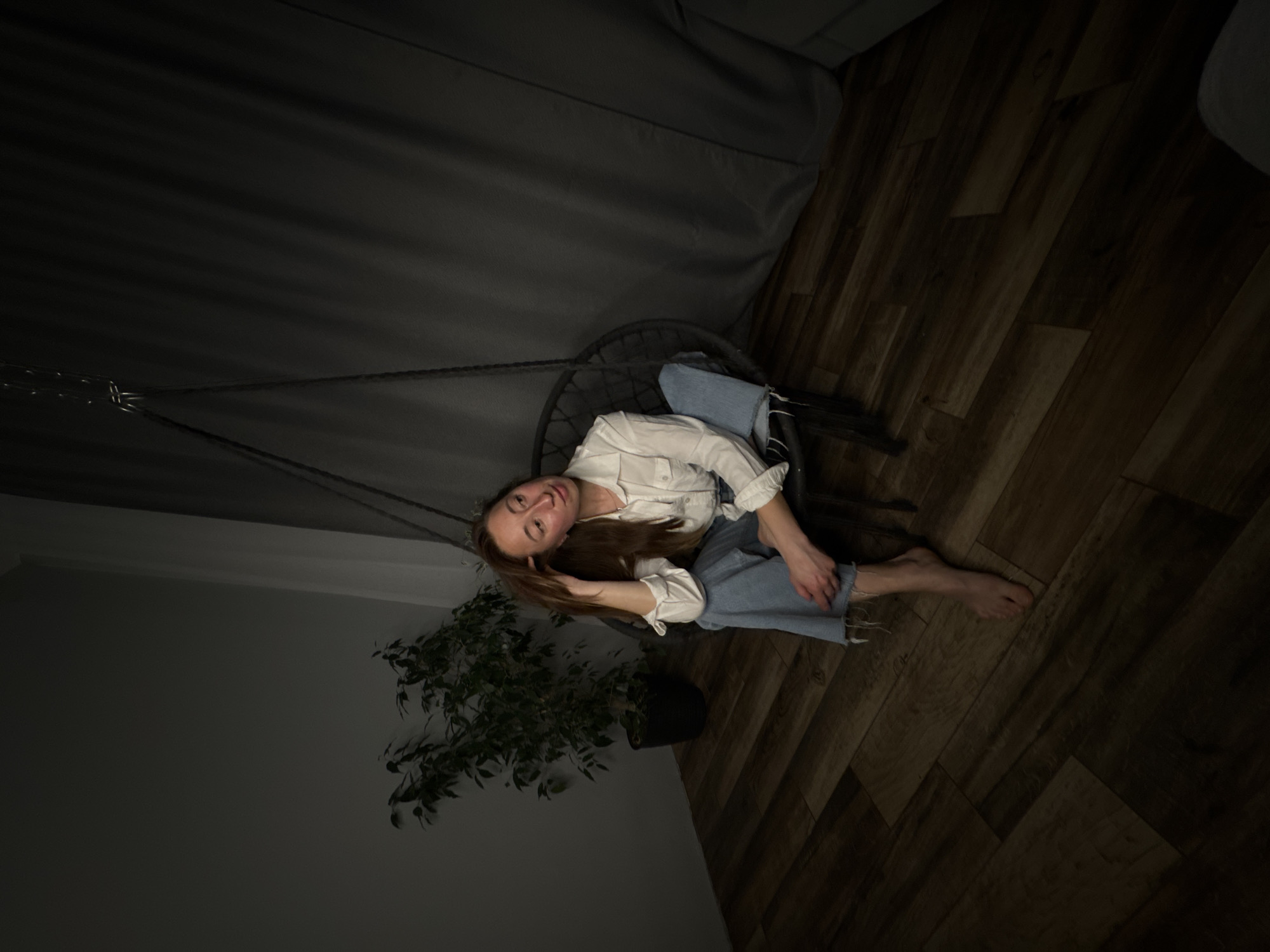 A high-angle indoor shot shows a woman seated in a hanging chair in a dimly lit room. The woman, with long brown hair, is wearing a white button-down shirt and blue jeans. She is barefoot, and her right hand is resting on her hair. The chair hangs from a cable attached to the ceiling. The room features dark hardwood floors, a grey curtain partially visible in the background, and a potted plant in the lower left corner. The image appears to have been taken in a location in the city of Chaikovsky, Russia.