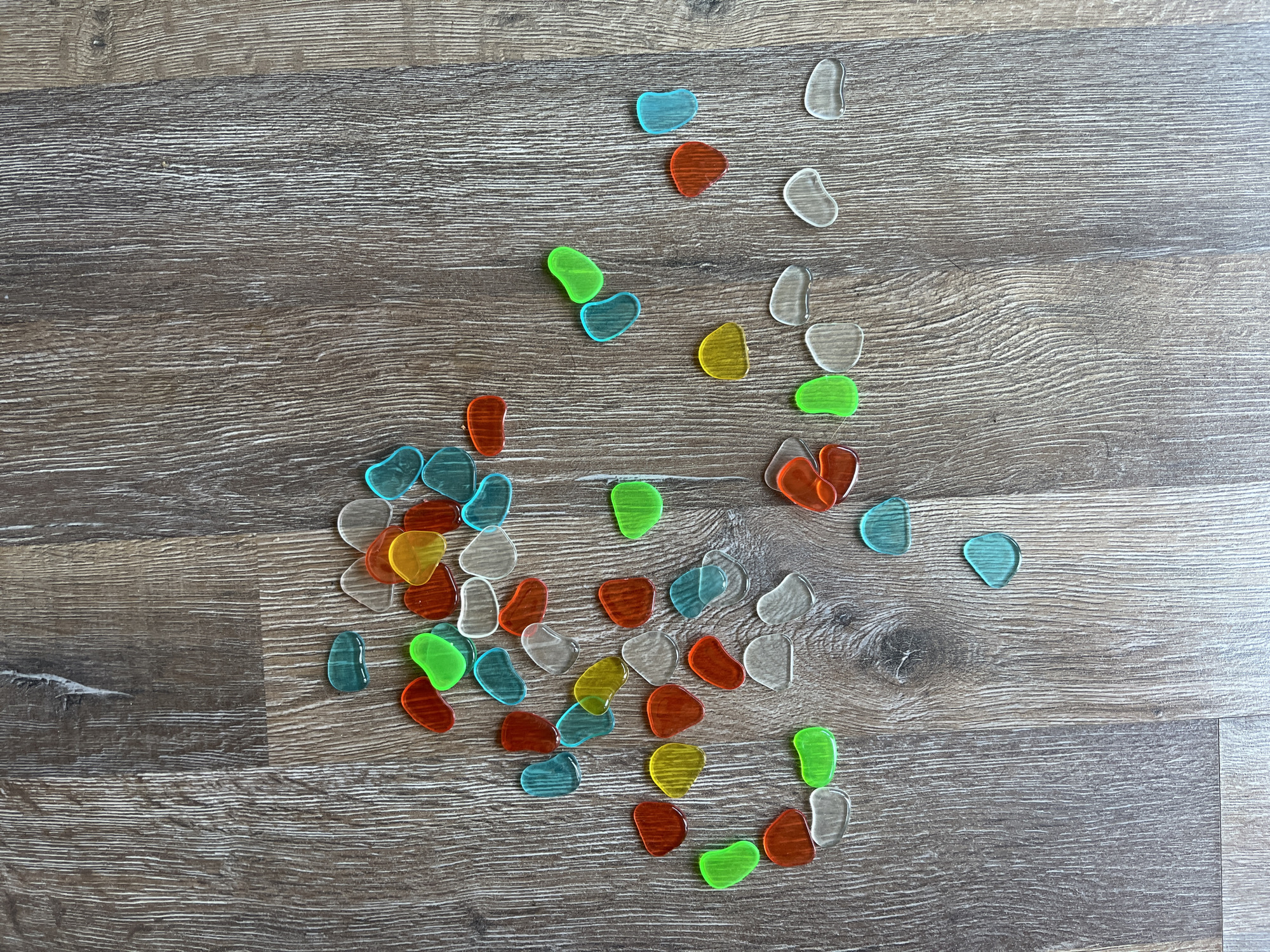 This image shows a collection of small, translucent, pebble-shaped objects scattered across a wooden surface. The objects are in various colors, including green, blue, yellow, orange, and clear. The wooden surface appears to be a floor or tabletop with a wood grain pattern. There is no indication of the city of Saratov, Russia, or any other specific location within the image. The scene is static and does not depict any actions or interactions.