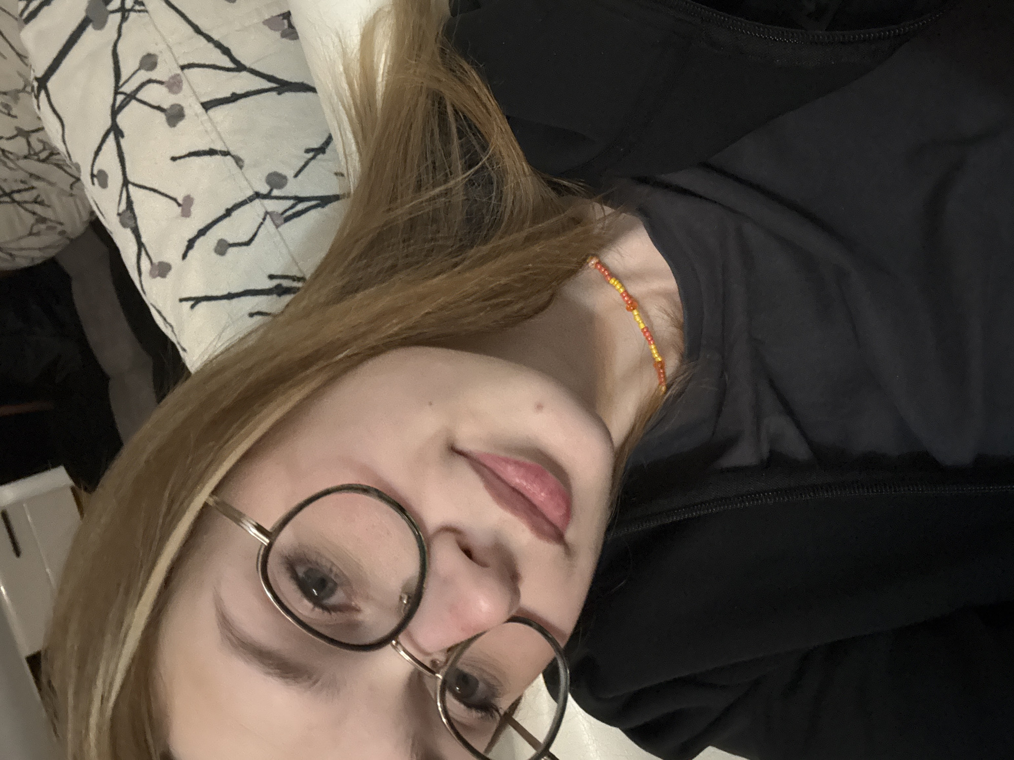 A close-up, rotated portrait shows a young woman with light brown hair, fair skin, and round-rimmed glasses, likely taking a selfie indoors. Her head is tilted to the right due to the image being rotated counter-clockwise. She is wearing a black top or jacket, and a thin, beaded necklace with red and yellow beads is visible on her neck. Her expression is neutral with a slight, soft smile, and she is looking directly at the camera. Her hair frames her face, with some strands falling over her shoulder. To the upper left of her head, a white pillow with a black and grey branching pattern is visible, suggesting she is lying down on a bed or sofa. The background is otherwise dark and indistinct. No specific location cues, weather information, or visible text are present.