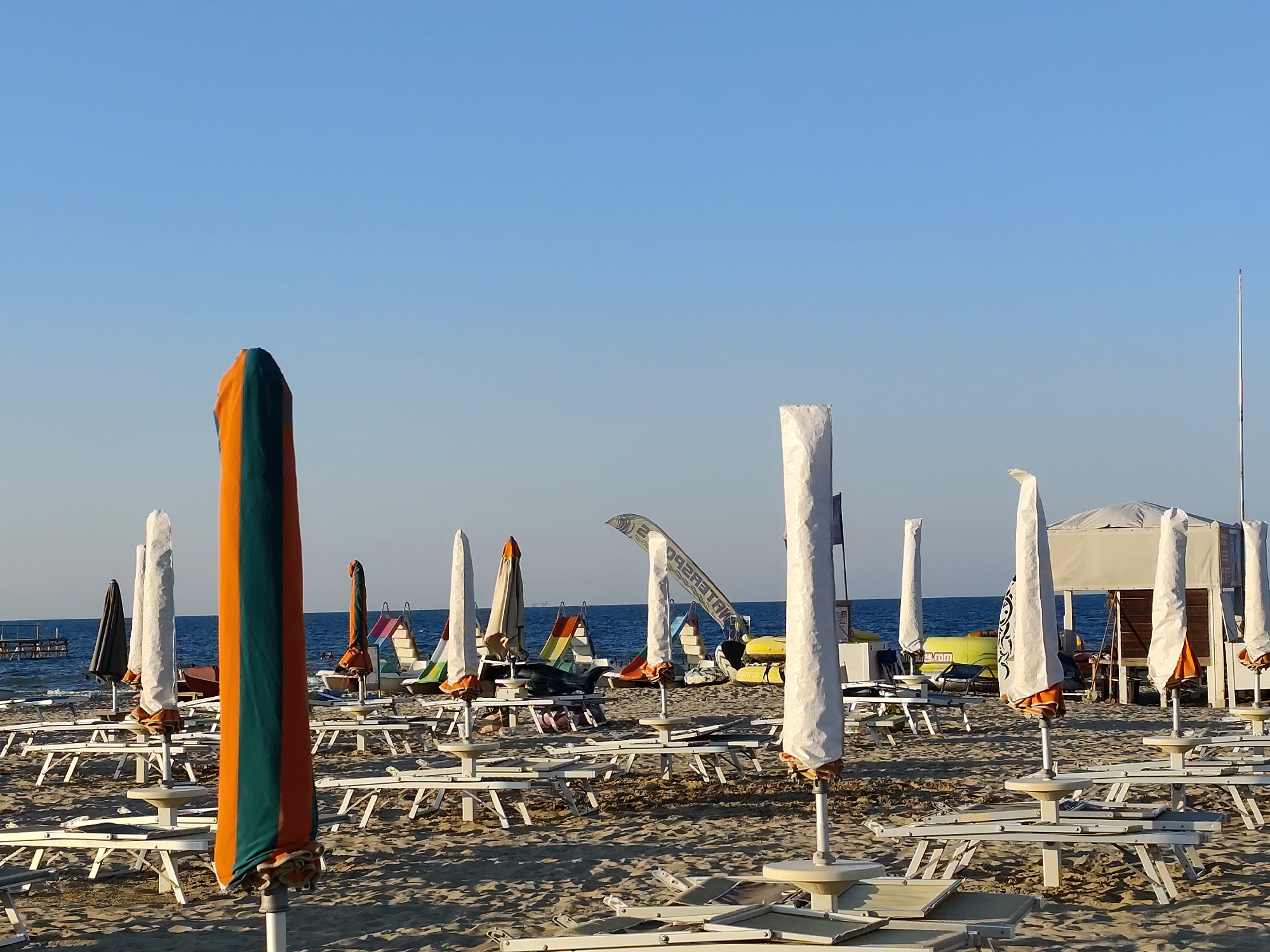 This is a photograph of an empty beach in Rimini, Italy. The image captures a wide view of the sandy shore, with rows of closed beach chairs and umbrellas. The umbrellas are mostly white and closed, with a few colorful ones visible in the background. Some of the chairs are stacked. The sea is visible in the background, with a clear blue sky above. There are no people in the immediate foreground, but some figures can be seen in the distance near the water. The scene appears to be set up for a day at the beach, but the umbrellas and chairs are closed, suggesting it is early morning or late evening, or that the beach is not currently in use.