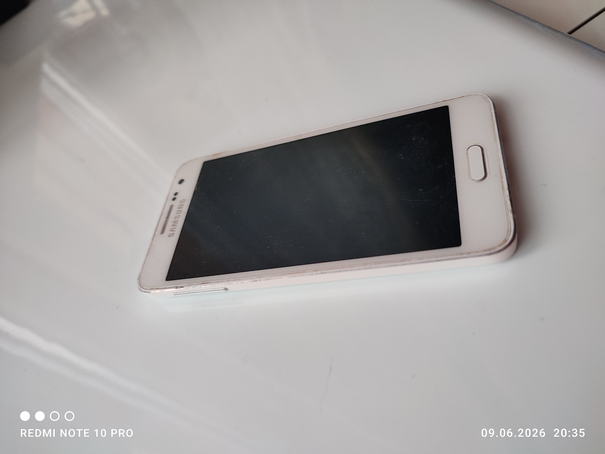 A white Samsung smartphone is positioned diagonally on a white surface. The screen is dark, and the phone shows signs of wear, with minor scuffs and smudges. The lower left corner of the image displays the text "REDMI NOTE 10 PRO" and a series of white dots. The lower right corner shows the date and time "09.06.2026 20:35". The background is a plain white surface with subtle reflections.