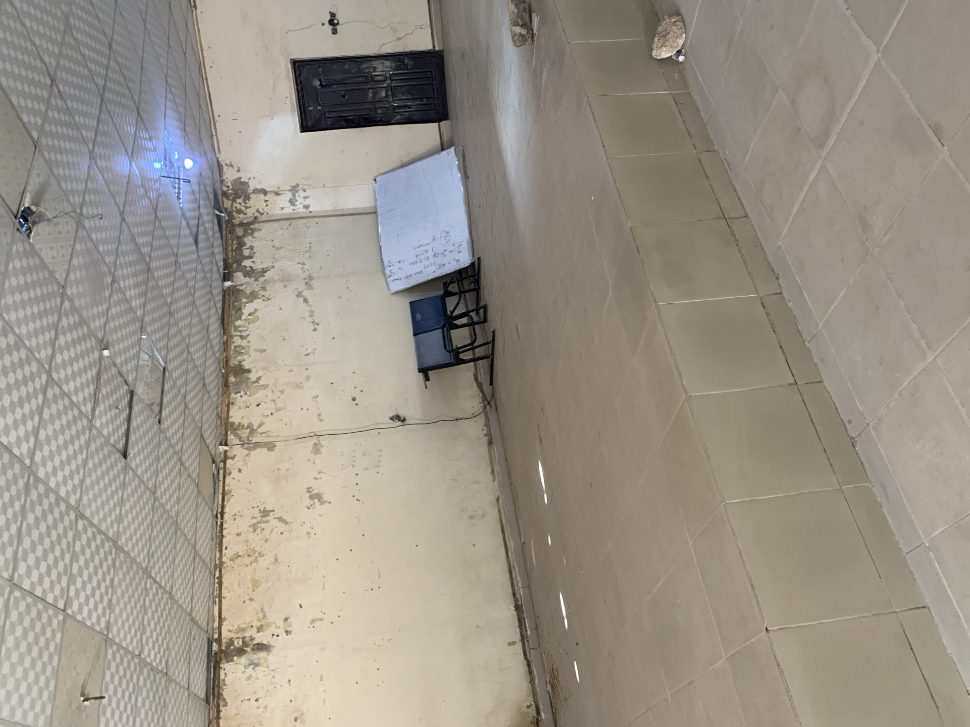 This is an image of an empty, dimly lit room in Jalingo, Nigeria. The room appears to be a former classroom or meeting space. The walls are unpainted and show signs of peeling plaster and water damage. The ceiling is a dropped ceiling with square tiles, some of which are missing or damaged. A single bare light bulb hangs from the ceiling, providing the primary illumination.
In the center of the room, there is a raised tiled platform, presumably a stage. On the stage, there are two blue chairs, a leaning whiteboard with some writing on it, and a closed black door. The floor is tiled with square beige tiles. There are also some rocks and debris scattered on the floor. The overall impression is one of disrepair and neglect.