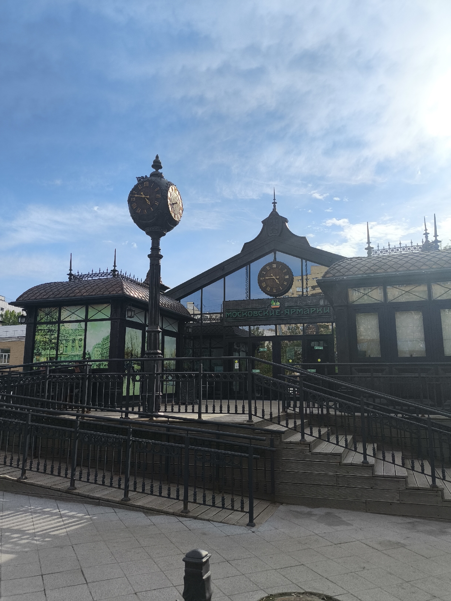 The image depicts the entrance to the Moscow Fair (Moskovskie Yarmarki) in Moscow, Russia. The building has a prominent clock mounted on its facade and a standalone clock tower in front. The architecture is ornate with dark metal framing and intricate details. Access to the entrance is provided by both stairs and a ramp, suggesting accessibility considerations. The sky is blue with scattered clouds, indicating a bright, sunny day. The foreground shows a paved area, likely a pedestrian zone. The overall scene is well-lit and appears to be taken during the daytime. No people are visible in the shot.