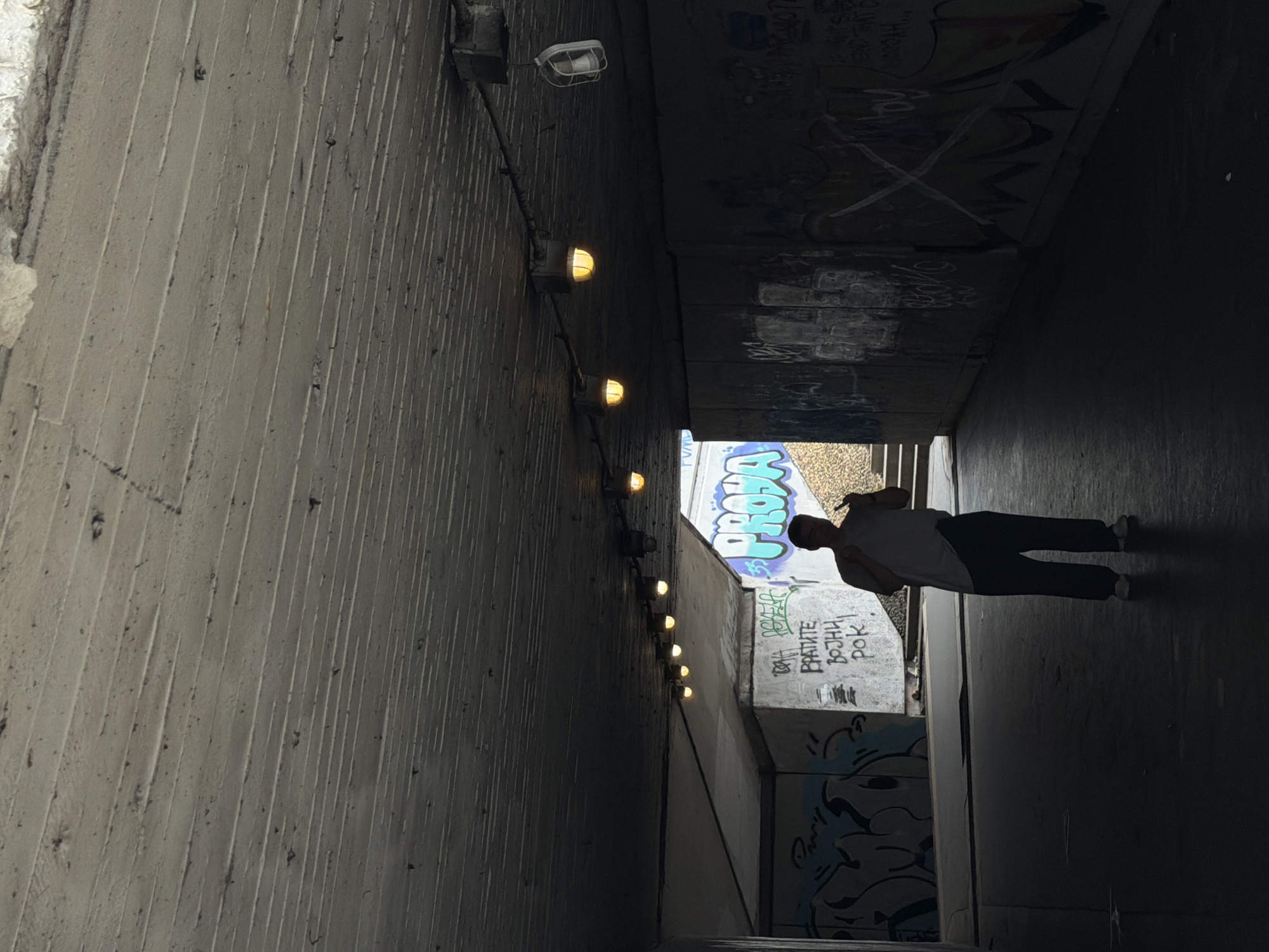 The image depicts an underpass in Belgrade, Serbia, illuminated by a series of evenly spaced wall-mounted lights. The walls of the underpass are concrete, with graffiti covering some sections. A person is walking in the underpass. The scene has a somewhat gritty, urban feel due to the graffiti and lighting.