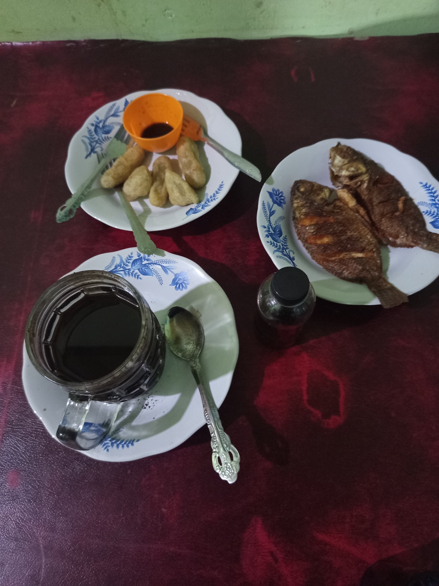 This image depicts a meal laid out on a dark red surface, likely a table. The meal consists of three plates. One plate holds two fried fish, appearing to be tilapia. Another plate contains several fried dough fritters and a small orange bowl filled with dark sauce. The third plate holds a glass mug filled with dark liquid, presumably coffee, and a spoon. A small dark-capped bottle sits next to the plate of fish. The plates have a blue floral pattern. The background suggests an indoor setting, with a green wall visible at the top. This type of simple, home-style meal with fried fish and coffee is common in Palembang, Indonesia.