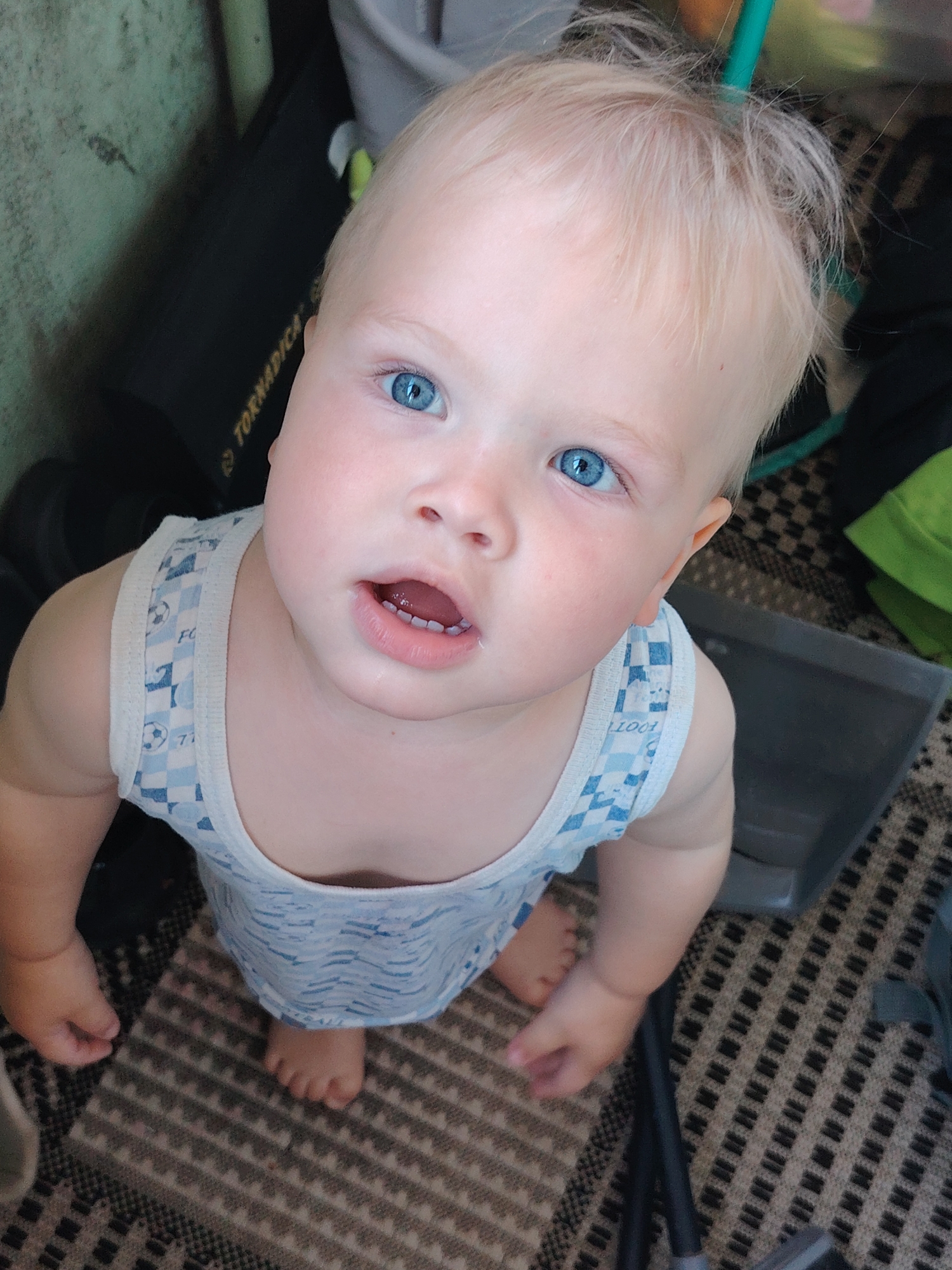 A baby with blonde hair and bright blue eyes looks upwards. The baby is wearing a light blue and white tank top with a checkered pattern and small soccer ball designs. The baby's feet are visible, and they are standing on a patterned rug. In the background, there is a black object with yellow text that appears to be a bag or case. There are also some dark gray and green objects, possibly containers or furniture. The overall scene appears to be an indoor setting. The image has been cropped and zoomed in to focus on the baby's face and upper body. This image was taken in Chaikovsky, Russia.