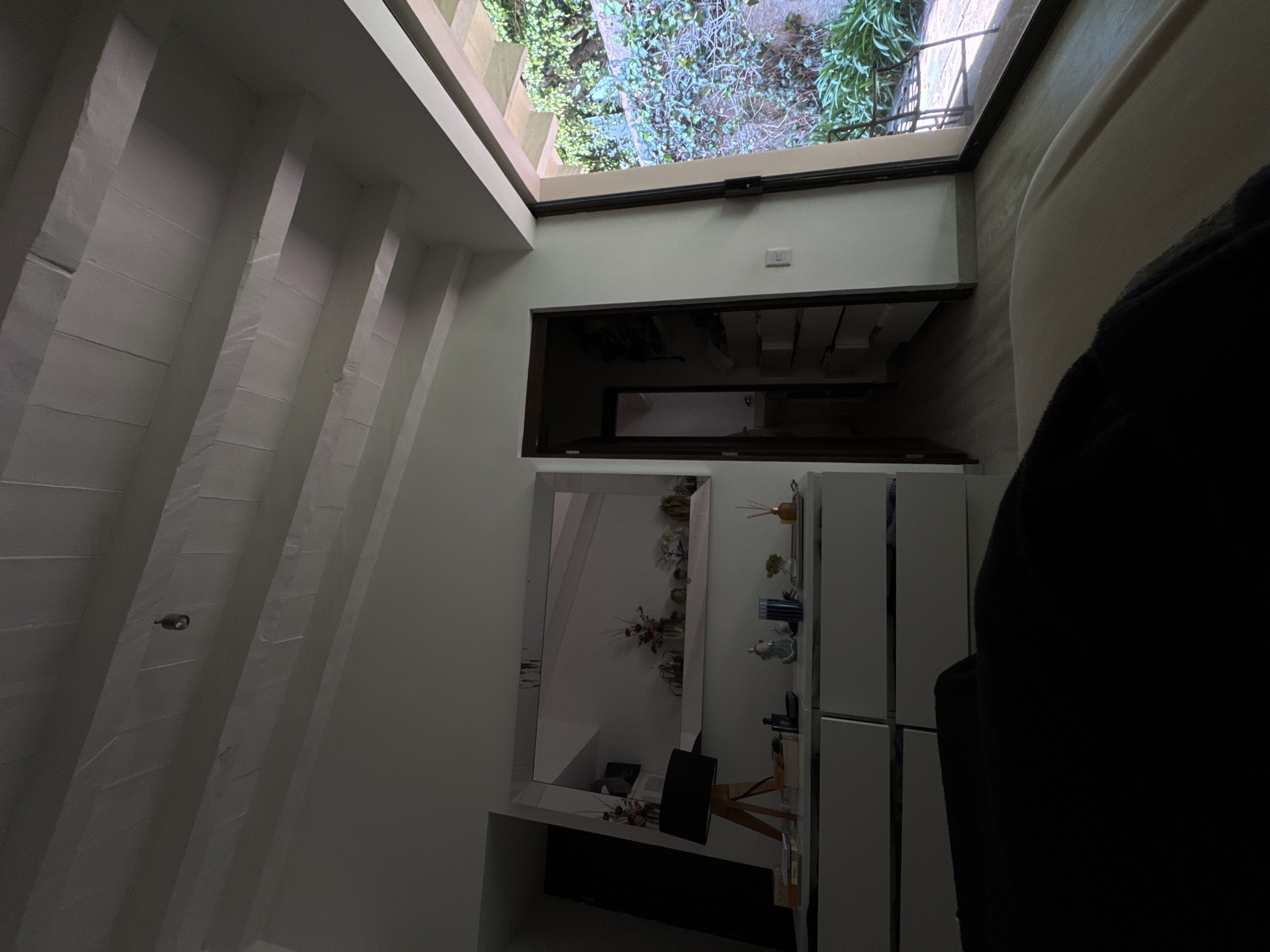 The image displays an interior space, possibly a hallway or transitional area, with a notable outdoor connection. The photo is rotated 90 degrees clockwise, so the description assumes a corrected vertical orientation.
On the left side of the frame, a tall, light-colored wall is characterized by several pronounced, regularly spaced vertical architectural features, resembling beams or pillars. A small, dark, cylindrical fixture, possibly a security camera or a light, is mounted on one of these vertical elements.
Towards the top, a large window or skylight offers a clear view of an outdoor scene. This exterior features abundant green foliage and plants, suggesting a garden or courtyard, with glimpses of a paved or stone ground and a metal railing. The natural light filtering through indicates it is daytime.
Below the outdoor view, the wall continues downwards. A rectangular, dark-framed opening or alcove is visible, appearing to be a storage unit or display area with shelves holding various items. A white light switch is mounted on the wall in this section.
Further to the right and lower, a large, rectangular mirror with a simple, clean design is mounted on the wall. Below the mirror, a wooden desk or console table is positioned, supporting a dark speaker and other decorative objects. Adjacent to this, a white multi-tiered cabinet or shelving unit displays an assortment of small items, including diffusers and decorative pieces.
A dark, blurry shape in the immediate foreground on the right suggests the shoulder and back of the person taking the photograph, providing a first-person perspective into the room. No discernible text is visible within the frame.