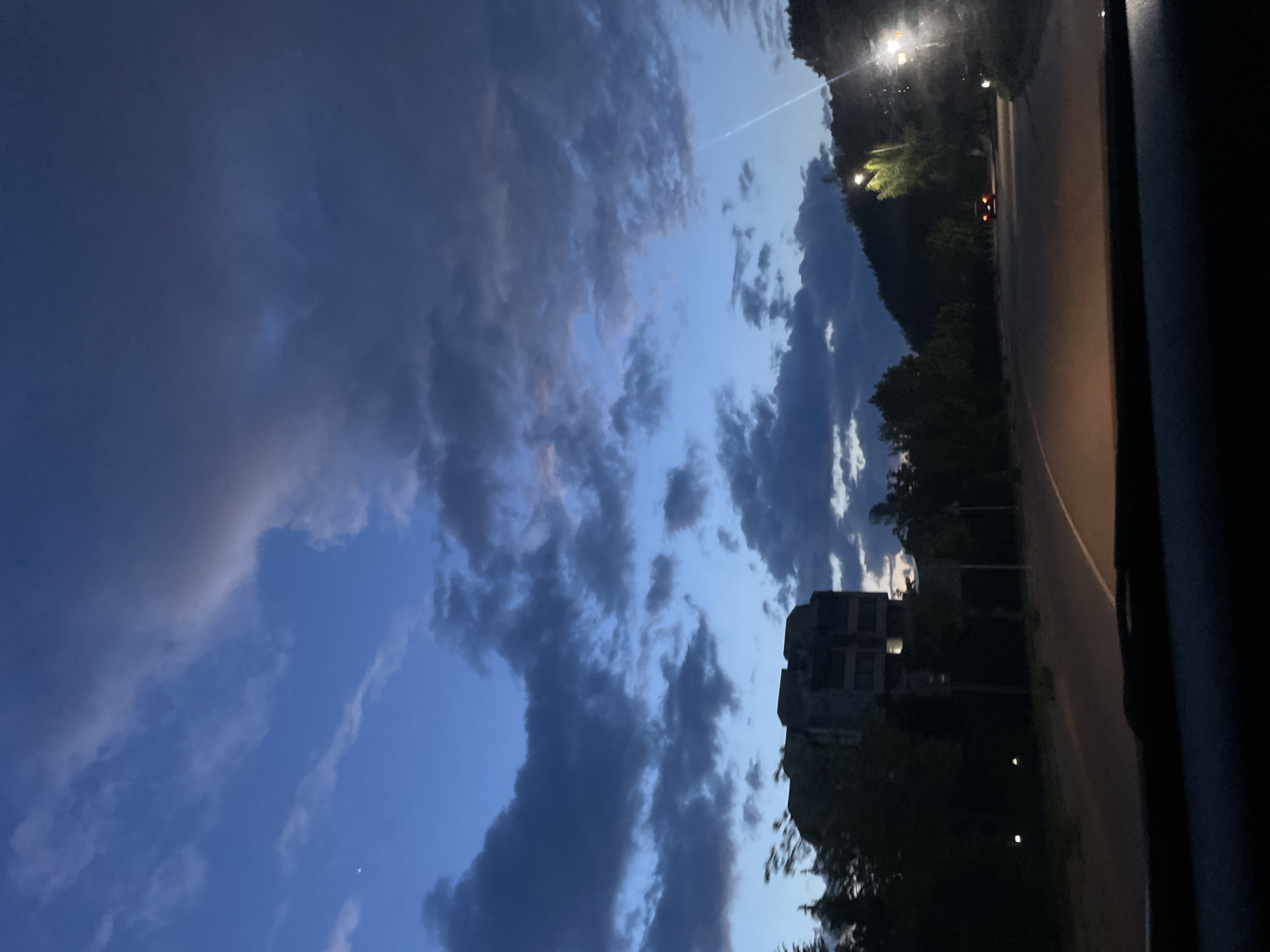 The image shows a view from a car at dusk/night in Zlatograd, Bulgaria. The car is driving on a road alongside trees and a building. The sky is cloudy with patches of light blue still visible. In the distance, there are street lights and possibly another car. The picture is taken at an angle.