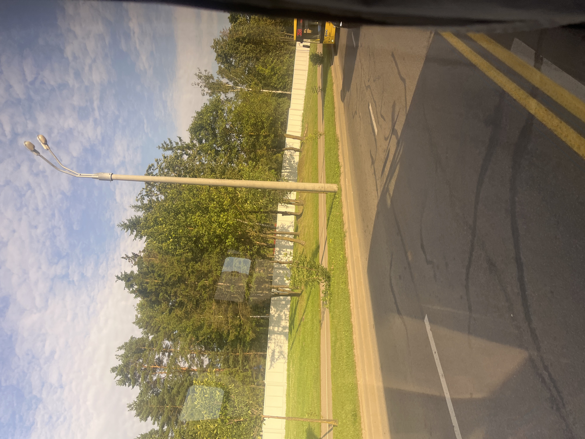 The image shows a street scene in Minsk, Belarus, on a sunny day with a partly cloudy sky. In the foreground, a paved road with yellow double lines and a white lane marker is visible. Shadows are cast across the road. To the left of the road, there is a grassy area with a sidewalk and young trees. Beyond that, a white fence runs along a line of trees, including pine and deciduous varieties. A tall lamppost with two lights stands near the road and trees. In the background, on the right side of the image, a yellow bus with the number 358 displayed on its front is driving on the road. The overall impression is a suburban or park-like setting within a city.