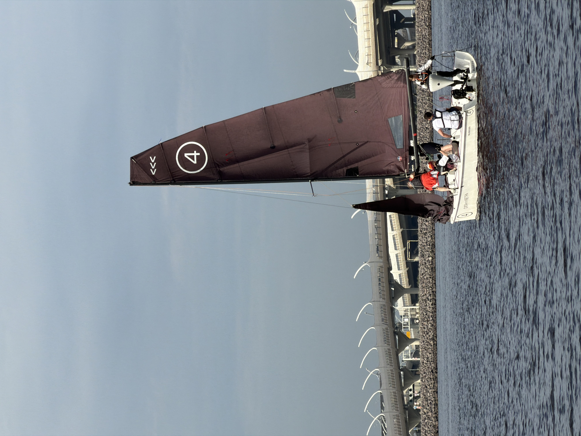 A white sailboat with a brown sail numbered "4" is sailing on dark blue water under a cloudy sky. The boat has at least four people on board, wearing life vests, with some appearing to be actively sailing. In the background, a modern elevated highway with white, curved barriers spans across the horizon, supported by concrete pillars. The highway structure and the style of the barriers suggest it is part of the Western High-Speed Diameter (WHSD) in Saint Petersburg, Russia, likely near the Gulf of Finland. The water is slightly choppy, indicating a moderate breeze. The overall scene depicts a recreational sailing activity with a prominent urban infrastructure element in the background.