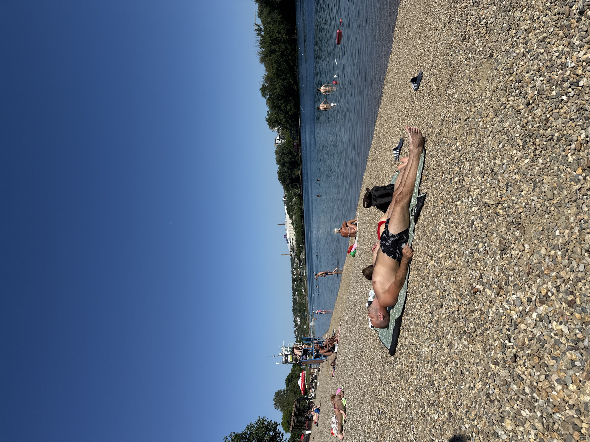 The image depicts a crowded beach on a sunny day, possibly Ada Ciganlija in Čukarica, Serbia. A man in swimwear lies on a towel on the pebble beach. Several people are swimming or standing in the water, while others are sunbathing on the beach. The beach also includes lifeguard stand. In the distance, there are trees and buildings along the shoreline.