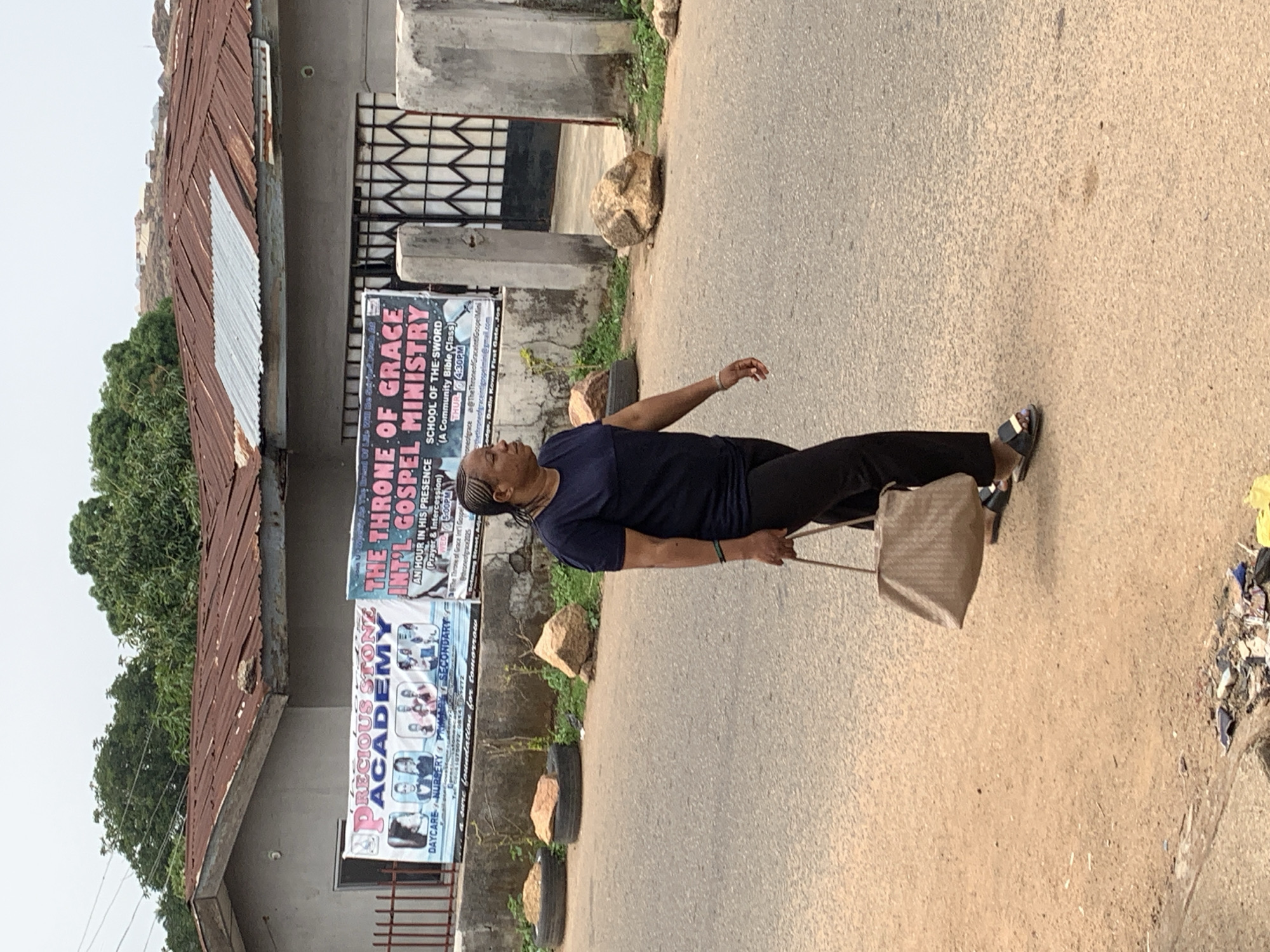 A middle-aged Black woman is walking on a paved road, carrying a light-colored handbag. She is wearing a dark blue t-shirt and black pants with sandals. The road runs parallel to a building with signs for "Precious Stone Academy" and "The Throne of Grace Int'l Gospel Ministry." The address on one sign is "Gomwalk Street, Jos," indicating the location as Jos, Nigeria. The building has a corrugated metal roof and a barred window. Debris is visible on the edge of the road. The woman is walking alone.