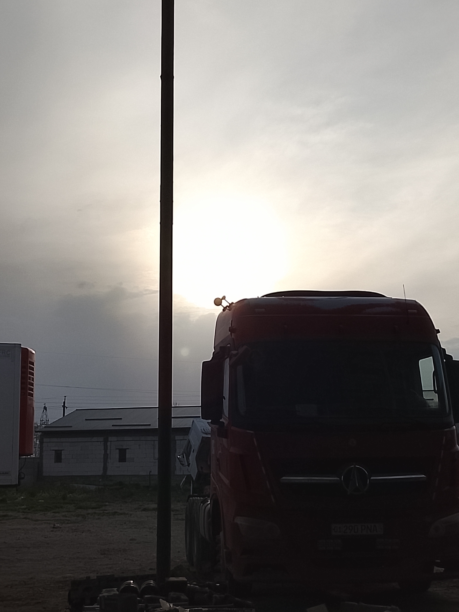 The image depicts a semi-truck in Sabir Rakhimov, Uzbekistan, potentially a Mercedes-Benz, parked on a dirt or gravel surface. The truck is positioned in the foreground and appears silhouetted due to the bright light in the background, making details difficult to discern. A pole is located to the left of the truck. In the background is a white, single-story building with a corrugated metal roof, and a white rectangular structure, possibly a refrigeration unit, on the left side of the image. The sky is overcast with the sun shining through the clouds. The license plate number "01 290 PNA" is visible on the front of the truck. There are miscellaneous objects at the bottom of the image, lying on the ground. No people are visible.
