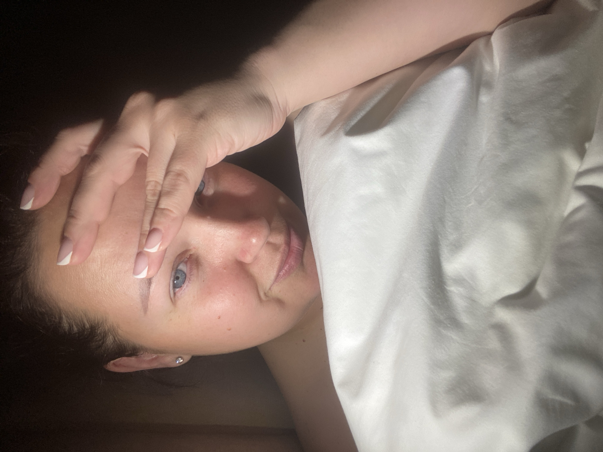 The image depicts a woman lying down, possibly in bed, with a white sheet partially covering her. She has fair skin, blue eyes, and dark hair. Her left hand is raised, with her fingers touching her forehead. The background is dark and indistinct. The location (Sochi, Russia) cannot be verified from the image.