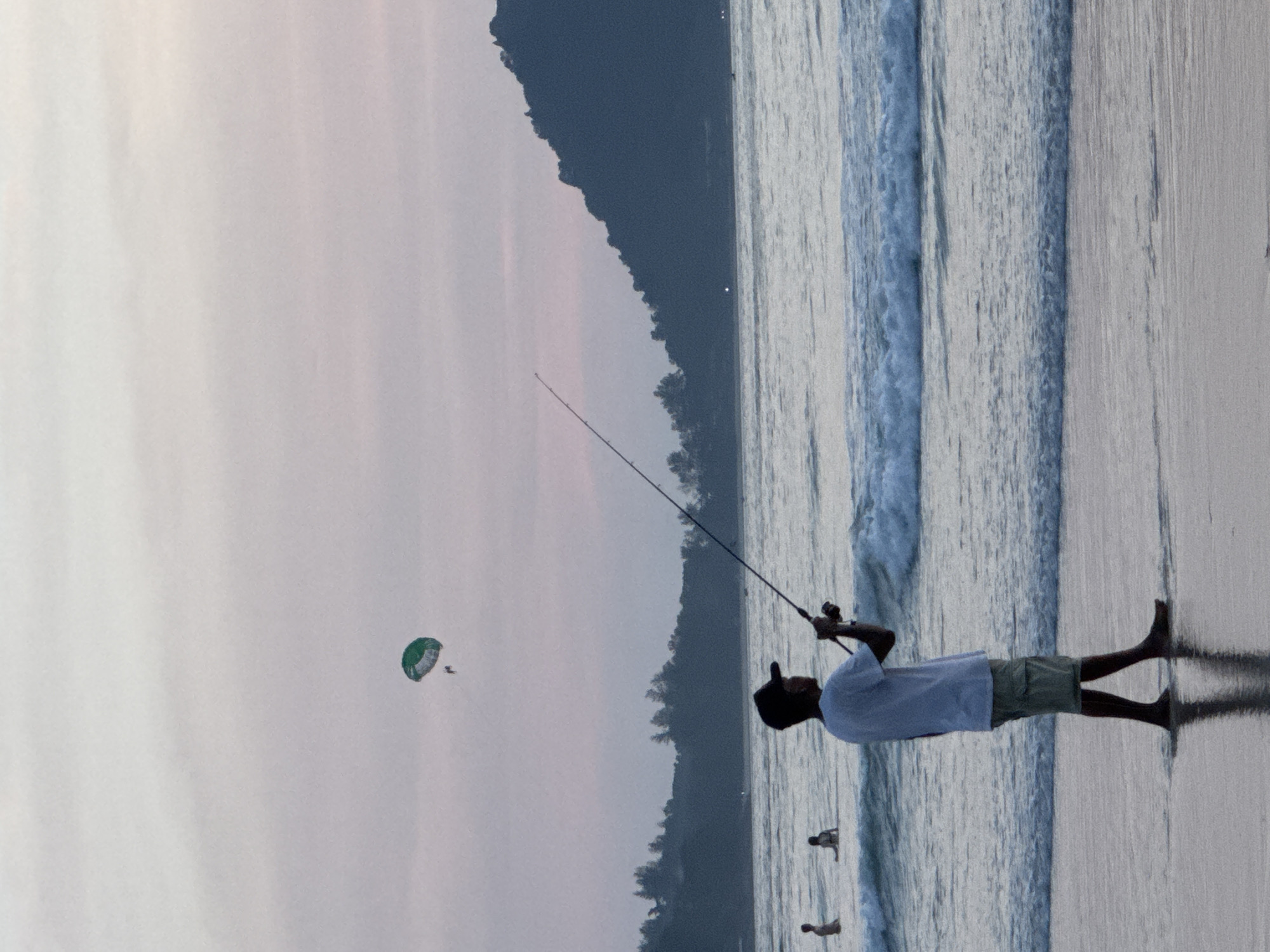 A young man, wearing a white t-shirt, khaki shorts, and a black baseball cap, stands in shallow water at the edge of a beach, holding a fishing rod with the line extended into the ocean. In the background, a parasail with green and white panels is visible in the sky, with two people suspended beneath it. The ocean has moderate waves, and a forested hill is silhouetted against a pale pink and gray sky. There are a few other people in the water further out. The scene appears to be on a beach in Patong, Thailand.