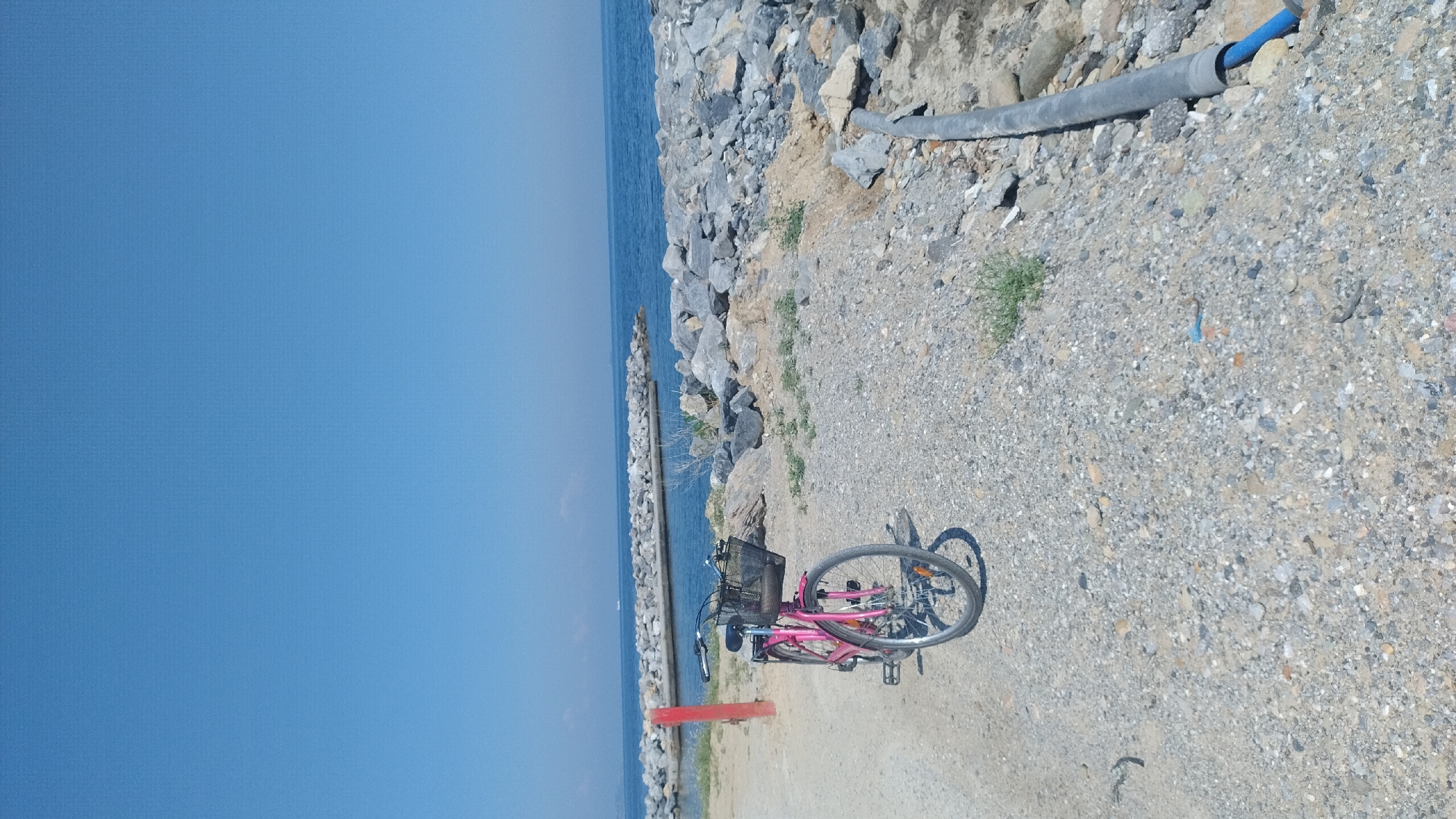 The image depicts an outdoor coastal scene in Thermi, Greece, under a clear blue sky. The top half of the frame is dominated by the unobstructed, vibrant blue sky. Below the sky, a calm blue body of water extends horizontally, with two grey rock breakwaters protruding into it from the right and center, creating a partially enclosed area. A faint white vessel is visible on the horizon line in the distant water to the left. The foreground and mid-ground consist of rough, light-colored gravel and small rocks, interspersed with sparse patches of dry green vegetation. A pink-framed bicycle with a black front basket and a black seat is parked on a dirt path in the left foreground, oriented slightly to the right, facing away from the viewer. Behind the bicycle, on the far left, a vertical red pole is embedded in the ground. In the bottom right corner, a long, dark grey pipe lies partially buried in the gravel, with a segment of blue pipe emerging from its end. No individuals are visible in the scene, and no overt actions or interactions are depicted. The environment appears dry and sunny.
