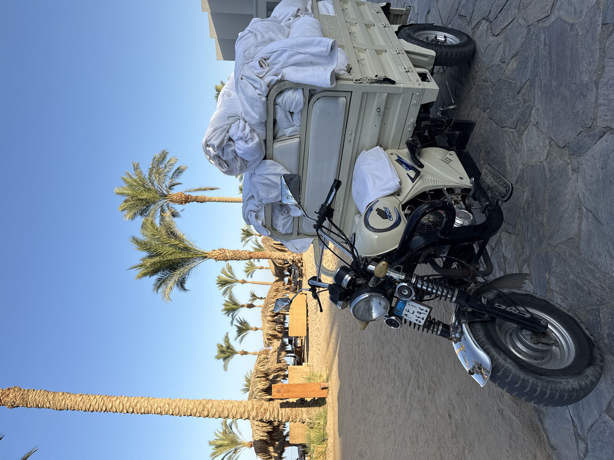 A motorized tricycle, painted a light cream color, is laden with a large pile of white towels, likely for a resort or hotel. The tricycle is parked on a stone pathway next to a sandy area with palm trees and thatched-roof sun loungers, suggesting a beachside or resort setting. The scene is set under a clear blue sky, indicating it's daytime with pleasant weather. The tricycle has a single headlight, a rear-view mirror, and a license plate with Arabic script, consistent with the location context of Hurghada, Egypt. The overall atmosphere appears to be one of preparation or service delivery in a tropical or subtropical tourist destination. The image is captured at an angle, with the tricycle positioned slightly off-center.
