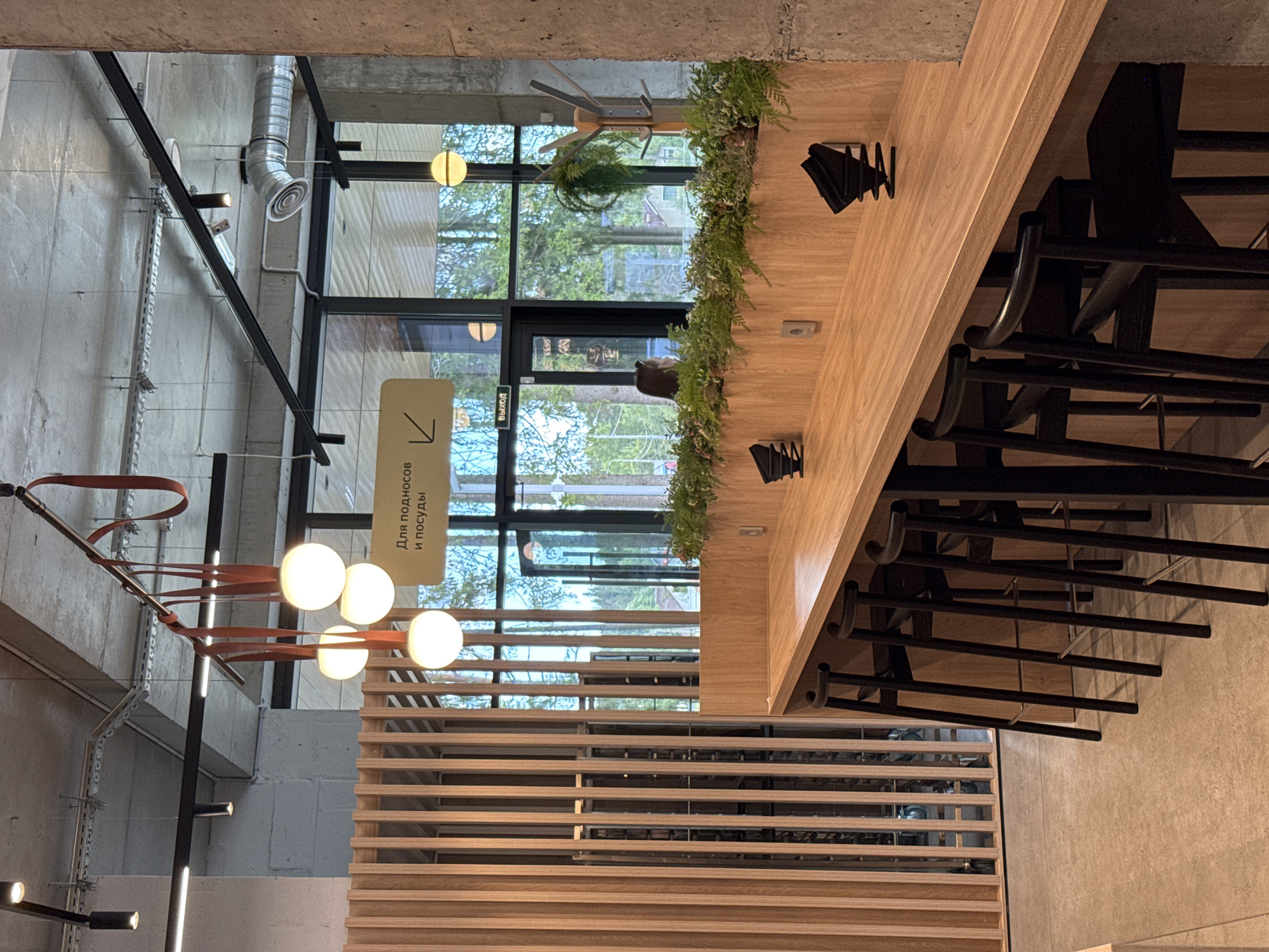 This is a wide shot of a modern cafe interior, possibly in Yakkima, Russia. A long, light-wood counter runs along the right side of the image, with several dark wooden bar stools lined up in front of it. Behind the counter, a large window overlooks a green, tree-filled outdoor area. To the left, a slatted wooden partition encloses a section of the cafe. Hanging from the industrial-style ceiling are multiple light fixtures, some globe-shaped and others cylindrical. A light-yellow sign with Russian text and an arrow is visible above the counter. A woman with dark hair is seated at the counter, partially obscured by plants. Several black decorative objects are placed on the counter.