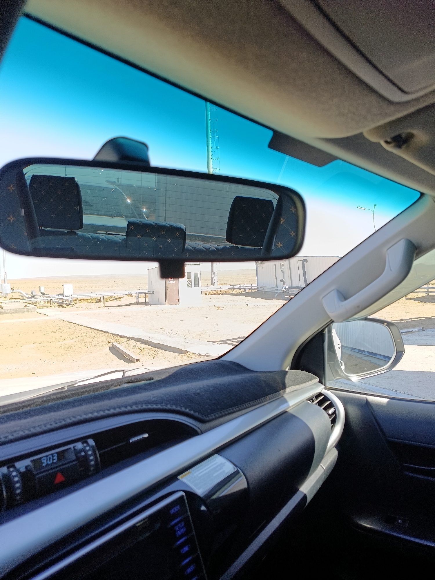 The image is taken from inside a vehicle, looking towards the windshield and side. The interior of the vehicle shows a black dashboard with a digital clock displaying "903", part of a car stereo, and a silver trim. A grey fabric dash mat covers the dashboard. The rearview mirror reflects the back seats, which have black upholstery with a subtle pattern, and the outdoor environment. The windshield is tinted blue, and the sky is clear and bright. Outside, a sandy, arid landscape is visible, with some industrial-looking structures, pipes, and a white building with a brown door. A street light is also visible in the distance. The overall scene suggests an industrial or remote location in a desert environment. No people are visible in the image. The location is identified as Unknown, Kazakhstan.