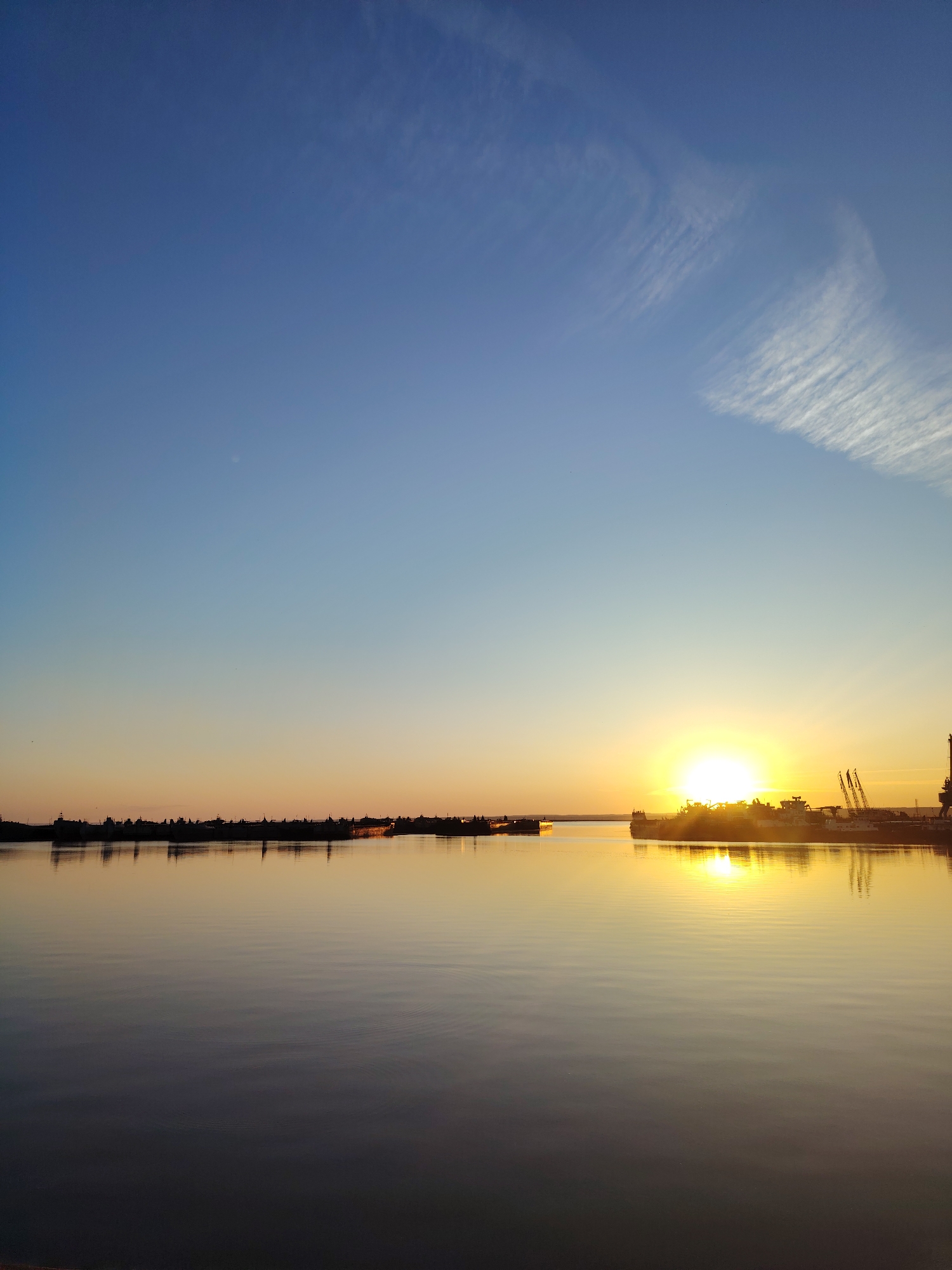 The image depicts a serene sunset over a body of water, likely the Kama River, in Chaikovsky, Russia. The sky transitions from a deep blue at the top to a soft orange and yellow gradient near the horizon. Wispy white clouds streak across the upper portion of the sky.
The water is calm, reflecting the colors of the sky and the silhouettes of distant structures. On the left side of the horizon, a line of dark, indistinct shapes, possibly buildings or industrial structures, is visible. To the right, a prominent silhouette of a ship or barge is positioned in the foreground, with several tall cranes rising behind it. The setting sun is a brilliant yellow disc, partially obscured by the ship, casting a strong, warm glow across the water's surface and the structures. The overall impression is one of tranquility and natural beauty at the close of day.