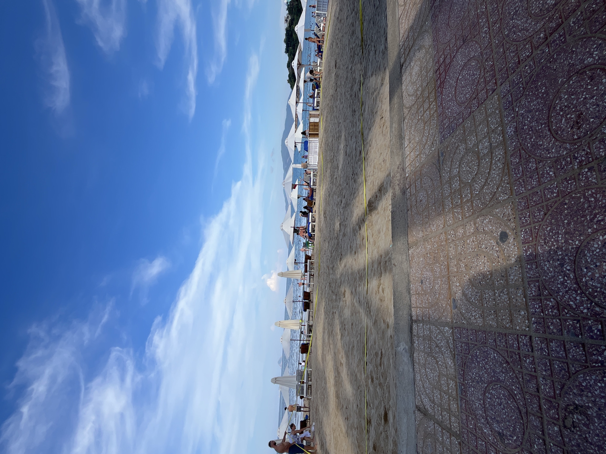 The image shows a beach in Bac Nha Trang Ward, Vietnam. White umbrellas and beach chairs line the sandy shore adjacent to a paved walkway. People are visible on the beach and in the water. The sky is blue with scattered white clouds. The paved walkway is patterned with alternating light and dark tiles.