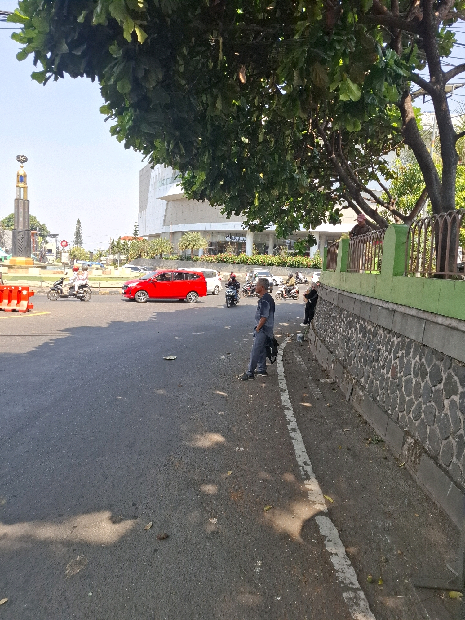 This is a street scene in Cianjur, Indonesia, on a sunny day. The foreground features a paved road with a white line marking the edge of a lane. Sunlight casts shadows on the road, and a few fallen leaves are scattered on the asphalt.
On the right side of the road, a stone wall topped with a green fence runs along the sidewalk. Two men are visible on the other side of the fence, one standing and looking out, the other leaning over the fence.
On the left side of the road, a man in a dark blue uniform stands on the sidewalk, facing away from the camera, with a backpack at his feet. Further down the street, traffic moves along. A bright red SUV is in the middle lane, and several motorcycles are also present. In the background, a large, modern building with a white facade stands under a clear blue sky. A monument with golden accents is visible to the left of the building. Trees with dense green foliage are prominent in the upper part of the image, casting shade. The environment suggests an urban setting with moderate traffic and pedestrian activity.