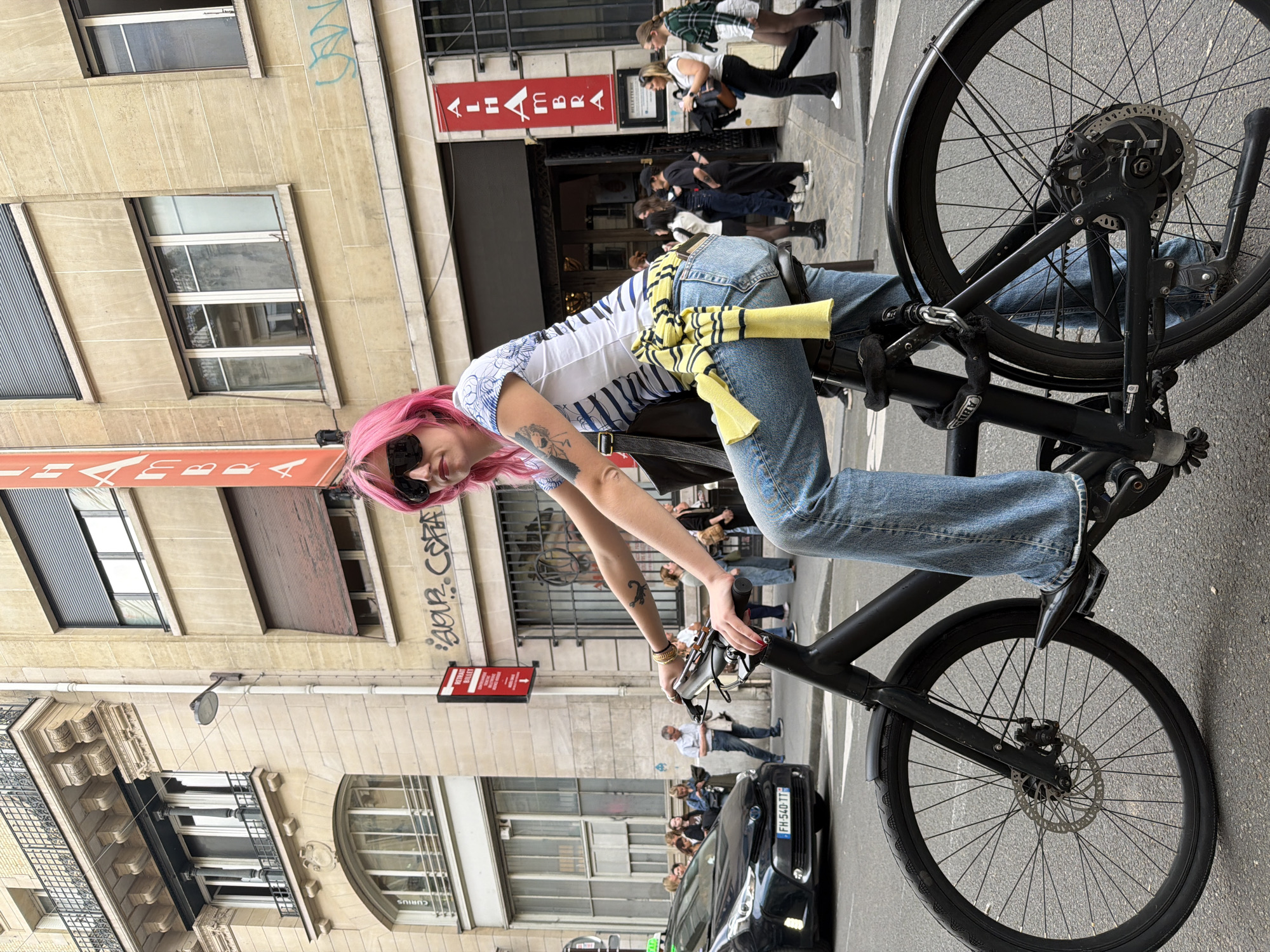 A medium shot of a woman with pink hair riding a black bicycle in Paris, France.
The woman has pink hair, sunglasses, a white and blue striped shirt, and blue jeans. A yellow sweater is tied around her waist. She is holding the handlebars of the bicycle and looking at the camera. Tattoos can be seen on her left arm. The bicycle is all black with disc brakes and thin tires. It is equipped with a rear-mounted electric motor.
The scene is set on a street in Paris. There are buildings on both sides of the street. Pedestrians are walking on the sidewalk. Cars are parked on the side of the road. Signage in the scene includes "ALHAMBRA BILLETTERIE", "CURIOUS" and graffiti reading "BOUR CERA". The license plate of the black car on the street reads "FH-540-TT".